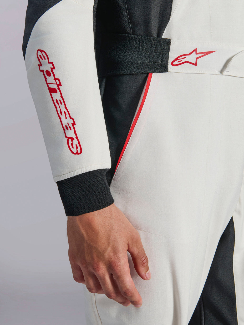 Tuta Alpinestars GP Tech V4, tuta da auto racing bianca, nera e rossa, primo piano della manica e della vita con marchio e logo a stella Alpinestars in rosso, vita elastica con chiusura a strappo e profili rossi su tessuto tecnico bianco e nero