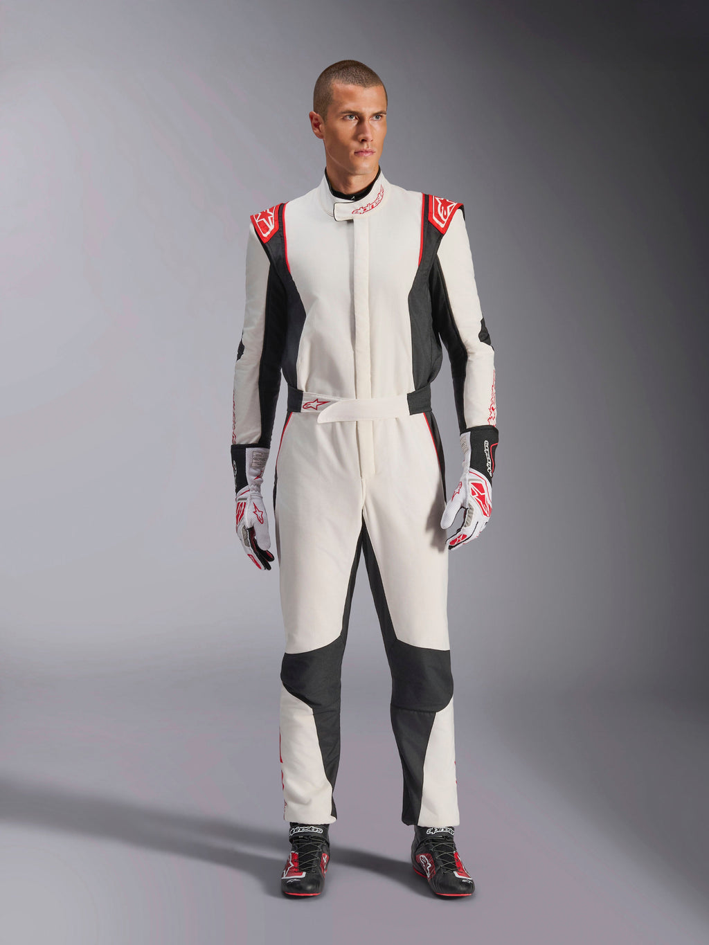 Tuta Alpinestars GP Tech V4, tuta da auto monopezzo bianca, nera e rossa, indossata da un modello con guanti e scarpe da corsa coordinati, con loghi rossi sulle spalle e pannelli laterali neri, abbigliamento professionale per motorsport per uso in pista