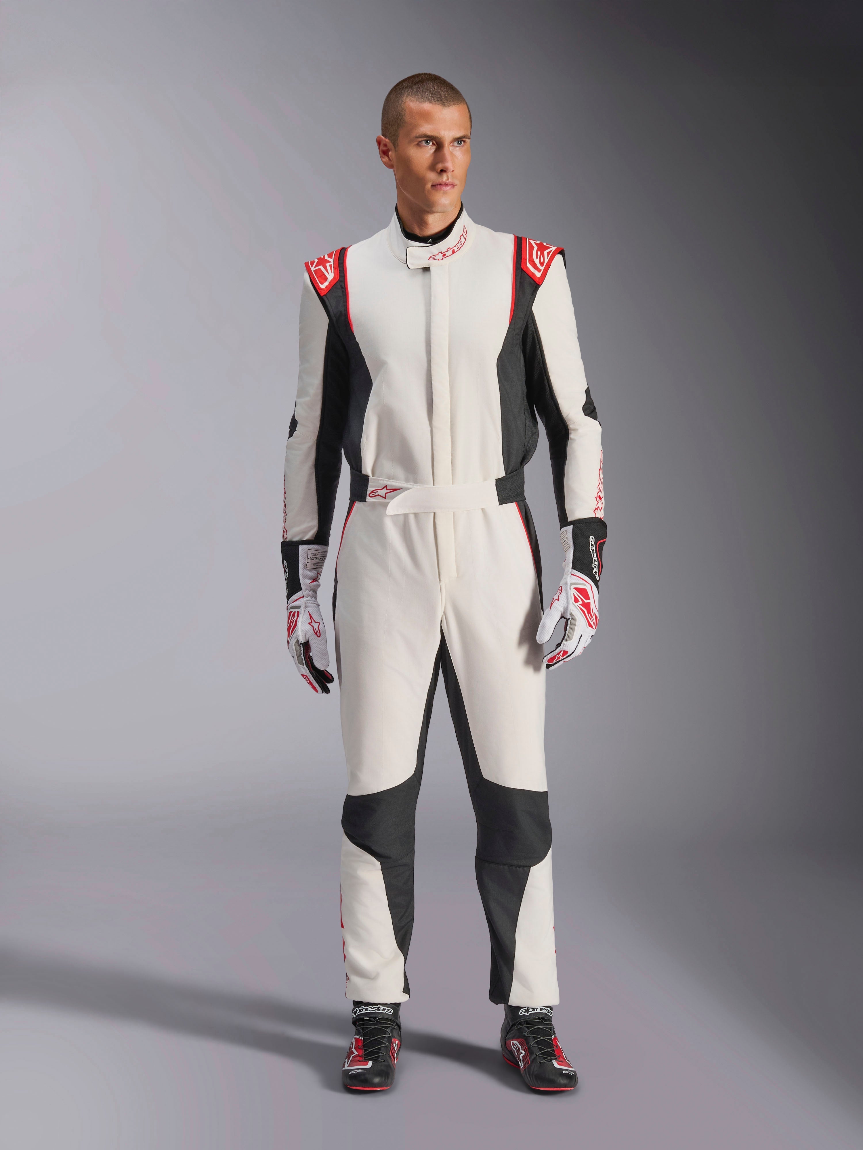 Tuta Alpinestars GP Tech V4, tuta da auto monopezzo bianca, nera e rossa, indossata da un modello con guanti e scarpe da corsa coordinati, con loghi rossi sulle spalle e pannelli laterali neri, abbigliamento professionale per motorsport per uso in pista