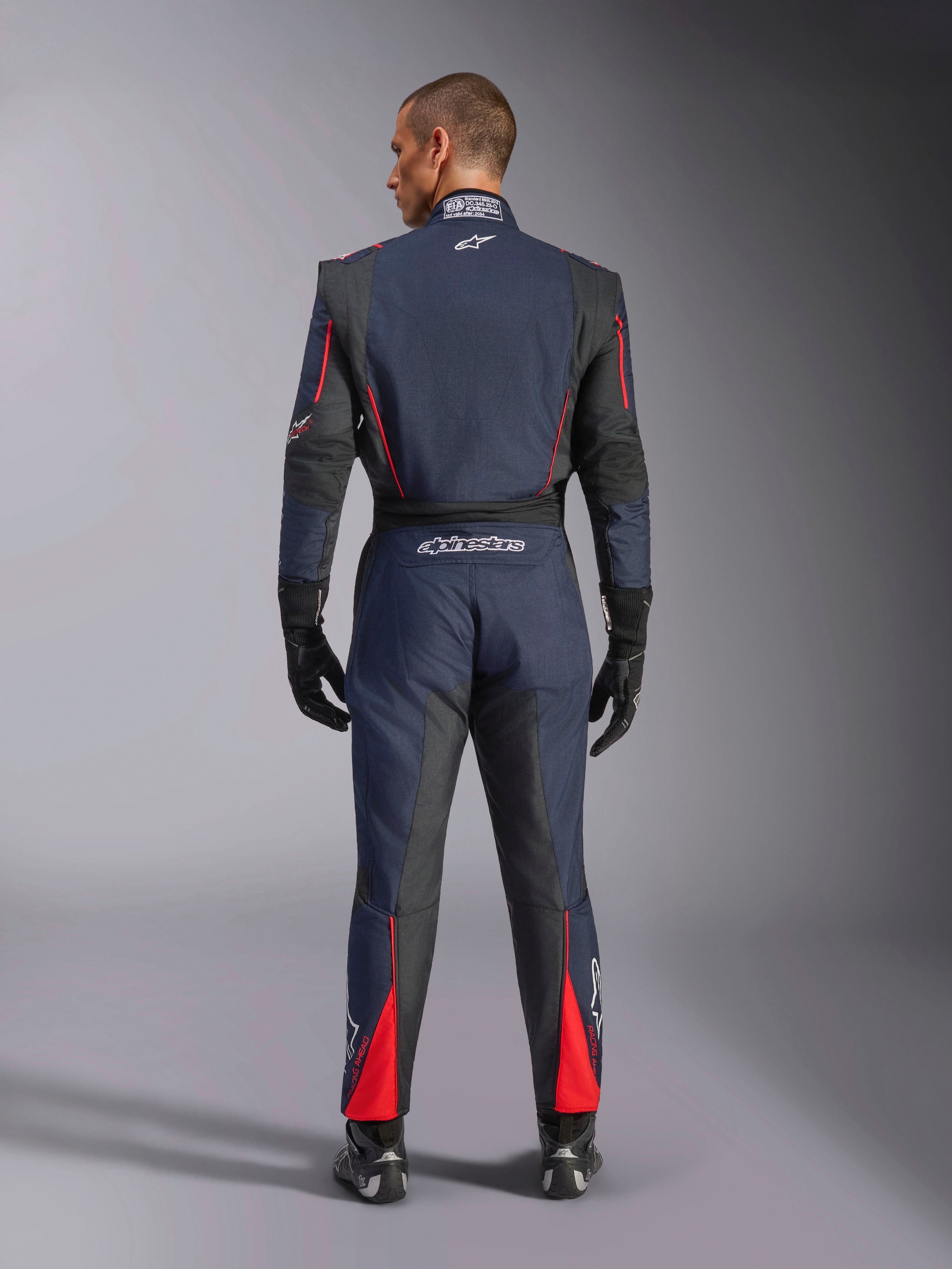 Vista posteriore della Tuta Alpinestars GP Tech V4 in Blu Navy Nero Rosso, tuta intera da corsa auto con pannelli elasticizzati neri e accenti rossi, marchio Alpinestars e dettagli di certificazione FIA, indossata con guanti e stivali da corsa professionali.