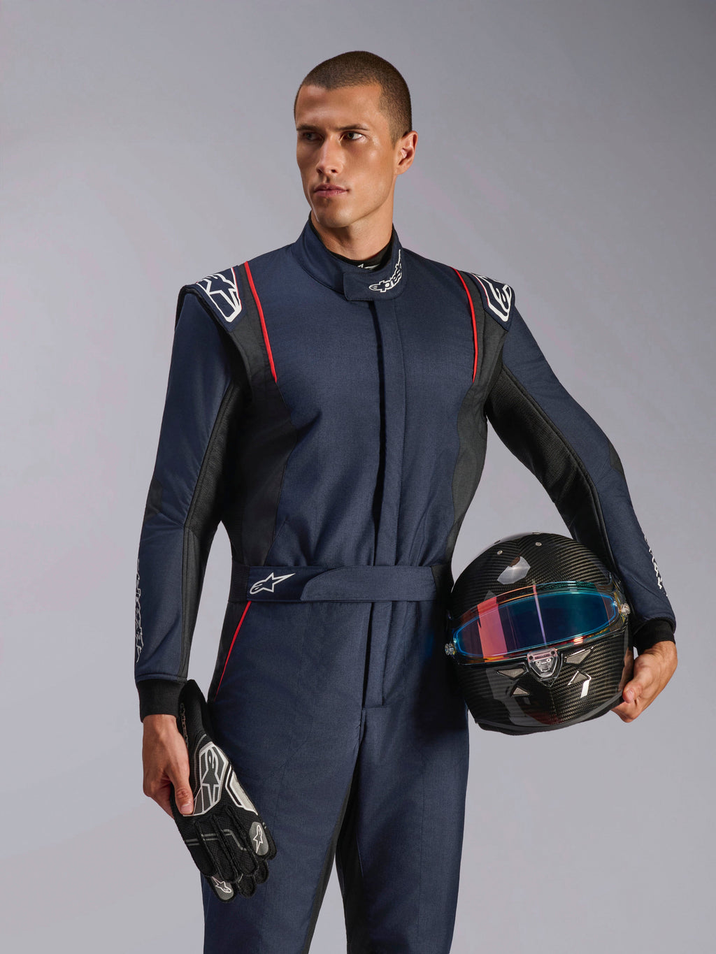 Modello che indossa la tuta Alpinestars GP Tech V4, tuta da auto racing blu navy con accenti neri e rossi, con in mano un casco in fibra di carbonio e guanti da corsa, abbigliamento protettivo professionale per motorsport