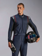 Modello che indossa la tuta Alpinestars GP Tech V4, tuta da auto racing blu navy con accenti neri e rossi, con in mano un casco in fibra di carbonio e guanti da corsa, abbigliamento protettivo professionale per motorsport
