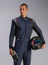 Modello che indossa la tuta Alpinestars GP Tech V4, tuta da auto racing blu navy con accenti neri e rossi, con in mano un casco in fibra di carbonio e guanti da corsa, abbigliamento protettivo professionale per motorsport