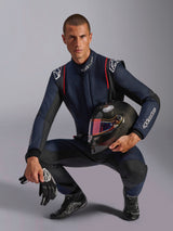Pilota automobilistico con tuta Alpinestars GP Tech V4, blu navy con accenti neri e rossi, regge un casco in fibra di carbonio con visiera iridescente, guanti e stivaletti da gara neri, abbigliamento professionale per competizioni automobilistiche