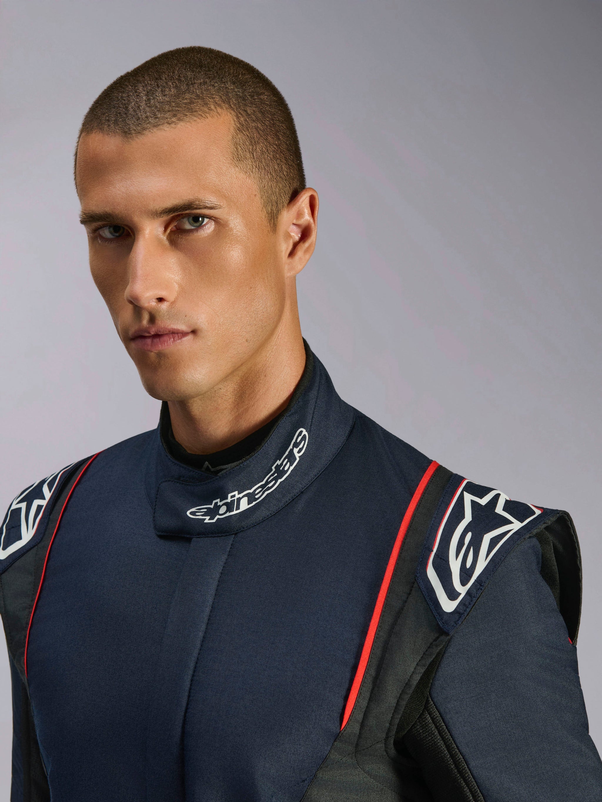 Ritratto in primo piano di un modello che indossa la tuta Alpinestars GP Tech V4, tuta da auto racing in blu navy, nero e rosso, con marchio Alpinestars sul colletto alto e spalline strutturate.