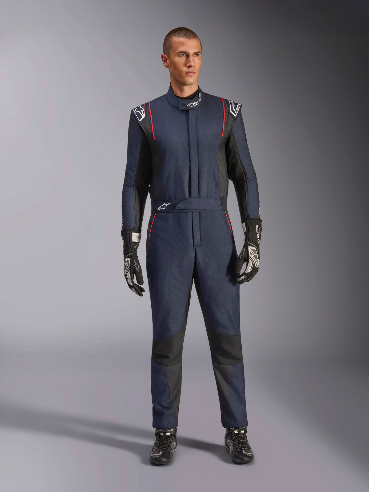 Tuta Alpinestars GP Tech V4, tuta professionale da auto racing in blu navy e nero con profili rossi, abbigliamento da corsa intero certificato FIA, indossata con guanti e calzature da corsa neri, abbigliamento per piloti di motorsport