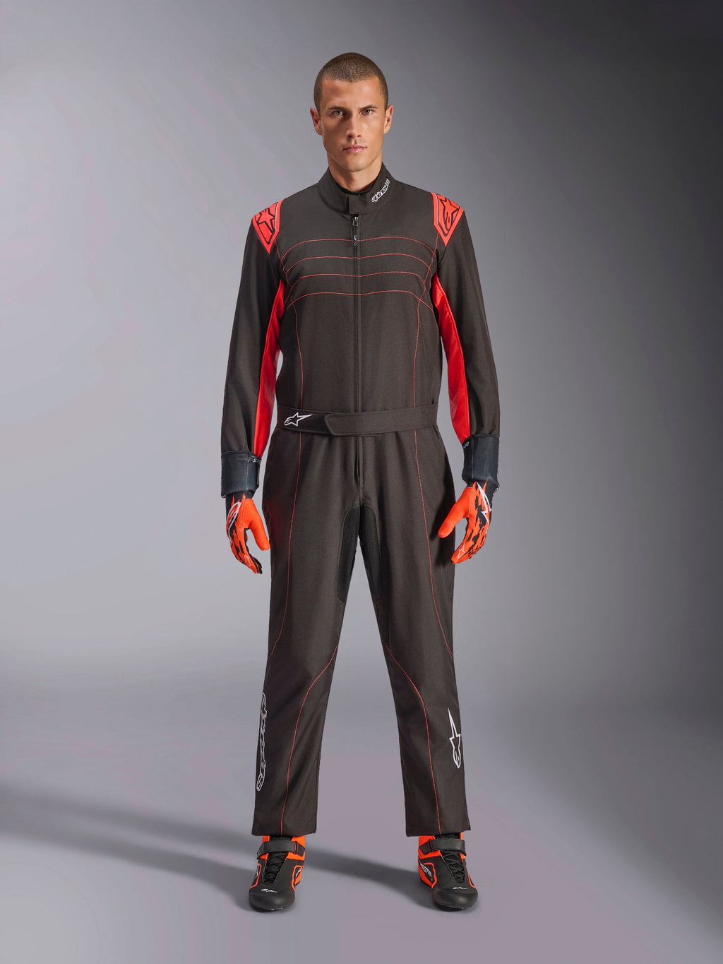 Tuta da Kart Alpinestars KMX-9 V3, nero e rosso fluo, tuta intera da corsa indossata con guanti e stivali coordinati, vivaci inserti rossi sulle spalle e cuciture, logo Alpinestars sulla parte inferiore delle gambe, design motorsport per corse in pista
