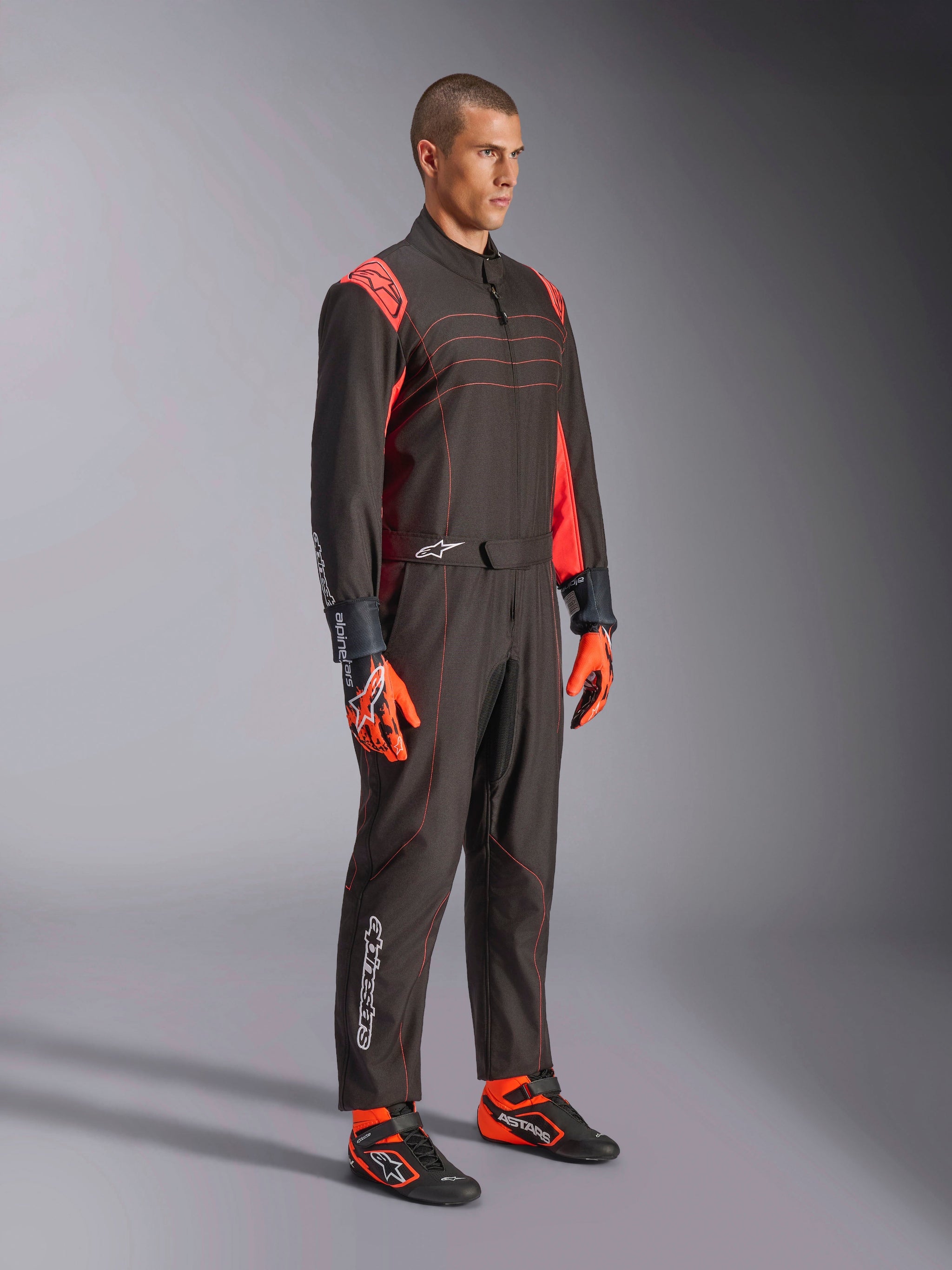 Alpinestars KMX-9 V3 Suit, Karting Suit, Black Red Fluo, worn by a model with orange gloves and black racing shoes, featuring red stitching, shoulder accents, and white logos, athletic design for professional racing
