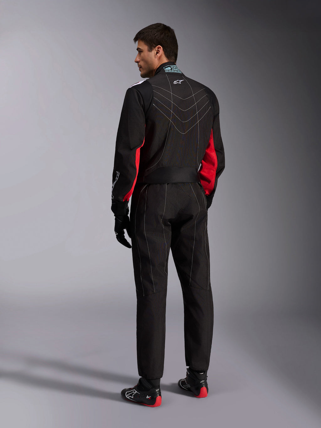 Tuta Alpinestars KMX-9 V3, Nero Rosso Bianco, tuta intera da karting vista dal retro, con cuciture geometriche bianche sulla schiena e dettagli rossi sulle maniche, indossata con guanti da corsa neri e scarpe da karting.