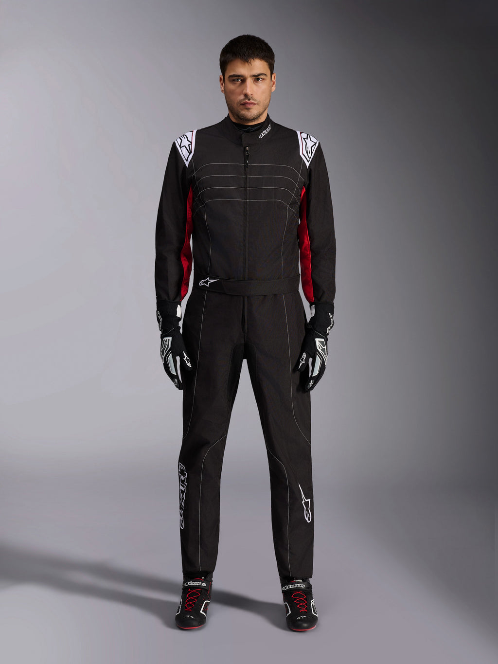 Tuta da kart Alpinestars KMX-9 V3 in nero, rosso e bianco, vista frontale a figura intera di un modello che indossa la tuta intera con guanti e scarpe da corsa coordinati, con loghi Alpinestars bianchi sulle spalle e dettagli rossi.