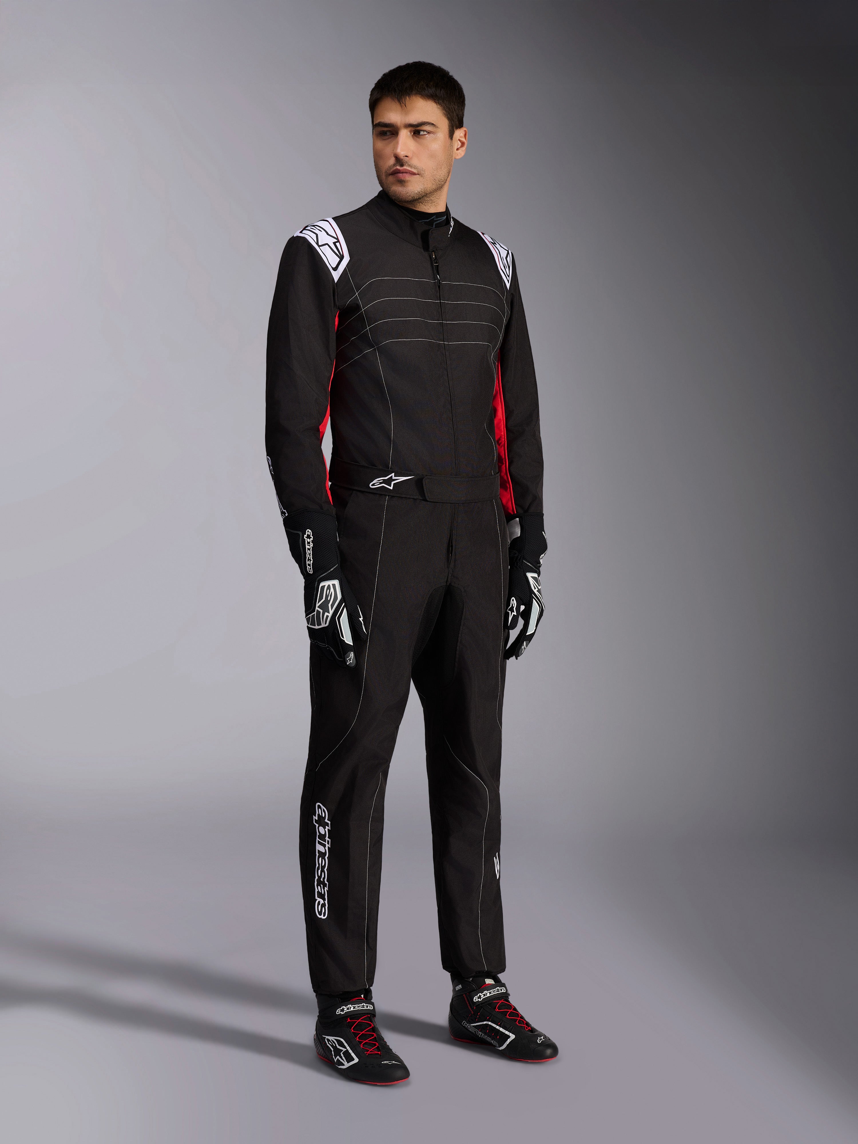 Tuta Alpinestars KMX-9 V3, tuta da kart nero rosso bianco indossata da un modello, con spalline e loghi bianchi, accenti laterali rossi, abbinata a guanti e scarpe da corsa coordinati, abbigliamento professionale per motorsport
