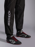Alpinestars KMX-9 V3 Suit, tuta da karting, nero e bianco, dettaglio della parte inferiore della gamba con grande marchio Alpinestars bianco, indossata con stivali da corsa neri e rossi, equipaggiamento professionale da motorsport per il karting