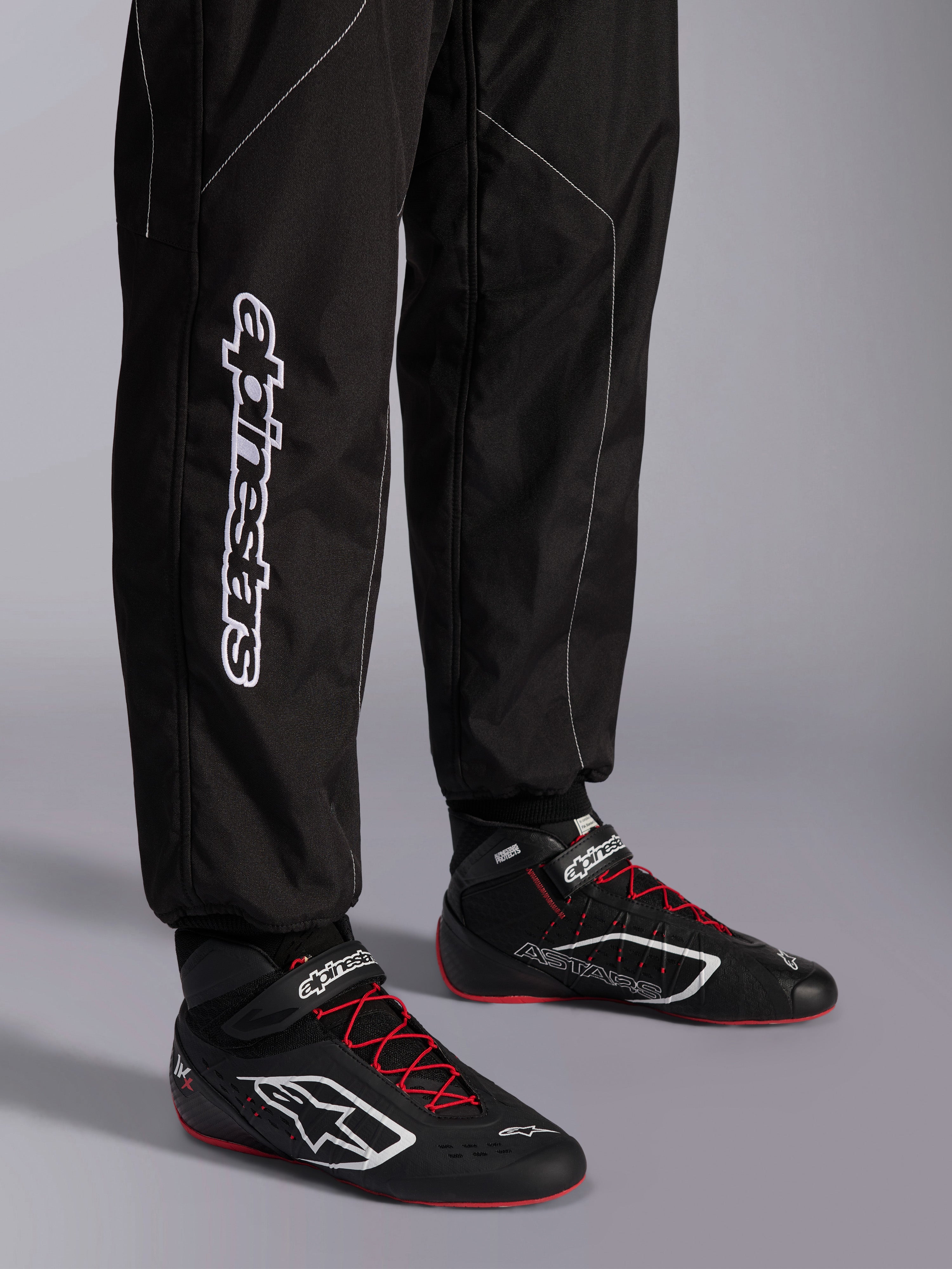 Alpinestars KMX-9 V3 Suit, tuta da karting, nero e bianco, dettaglio della parte inferiore della gamba con grande marchio Alpinestars bianco, indossata con stivali da corsa neri e rossi, equipaggiamento professionale da motorsport per il karting