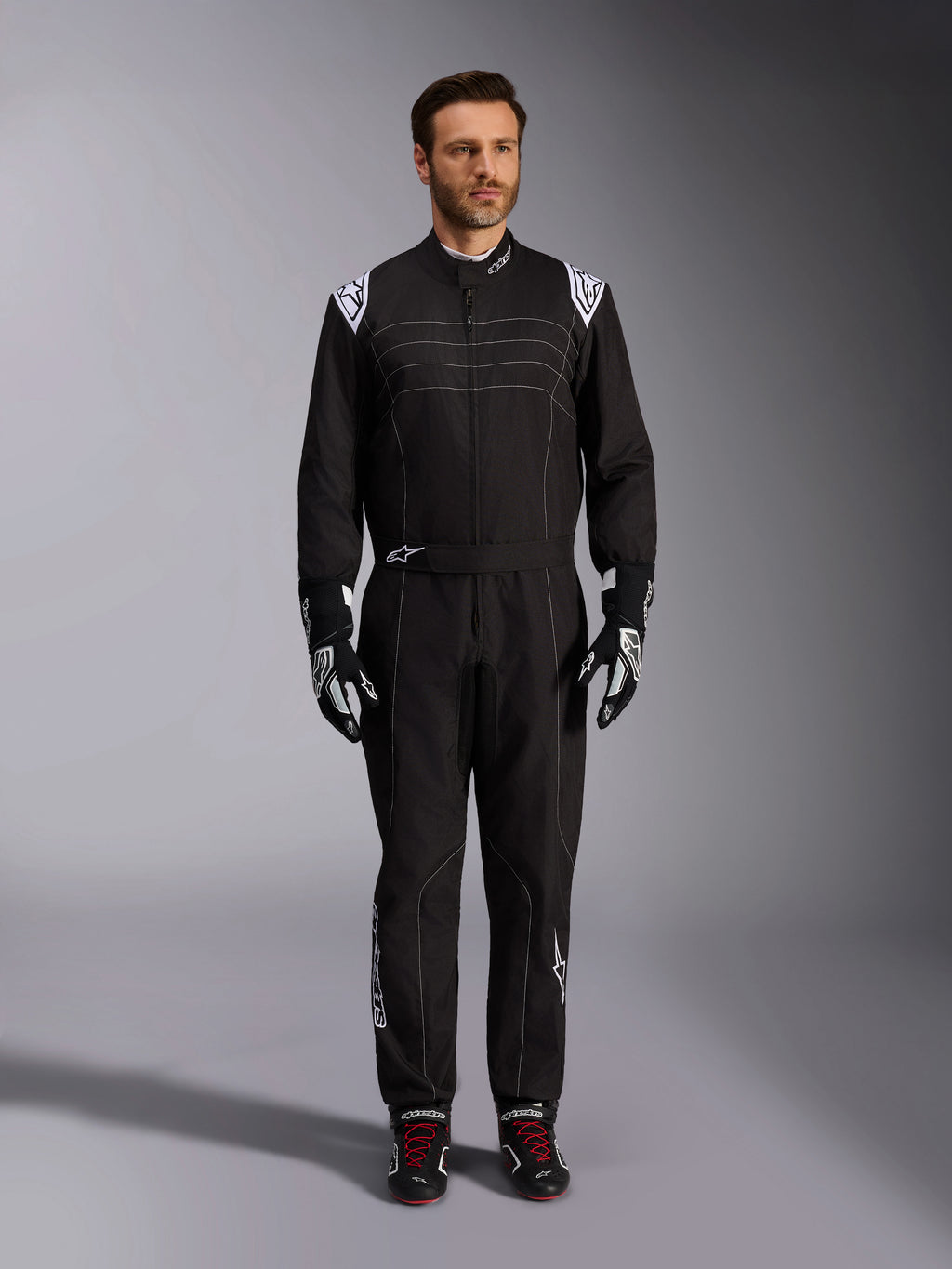 Tuta Alpinestars KMX-9 V3, tuta da karting nera e bianca, vista a figura intera su un modello, con loghi e dettagli bianchi sulle spalle, abbinata a guanti da corsa bianchi e neri e scarpe nere con lacci rossi, vestibilità athletic-regular per abbigliamento da karting professionale.