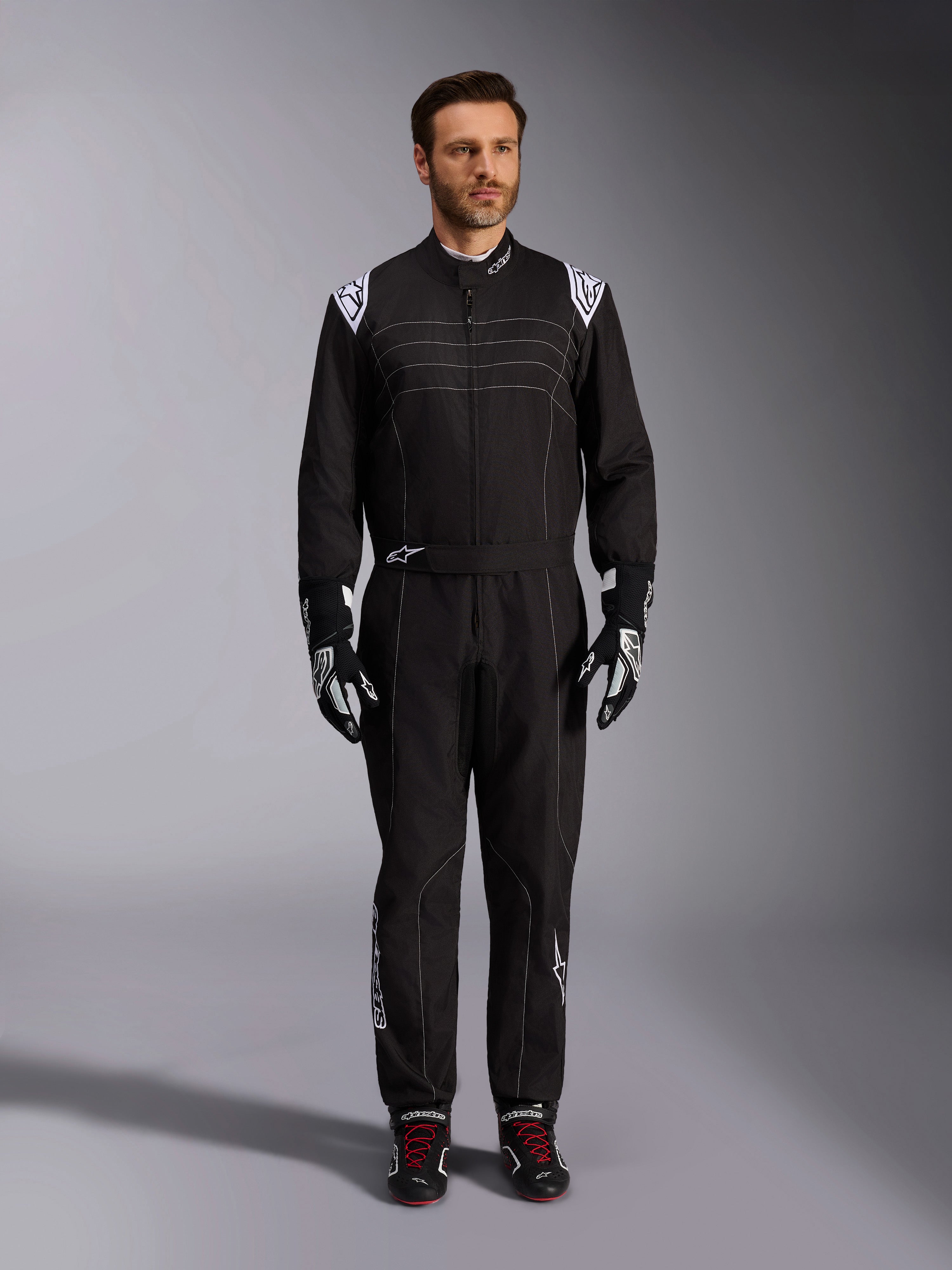 Tuta Alpinestars KMX-9 V3, tuta da karting nera e bianca, vista a figura intera su un modello, con loghi e dettagli bianchi sulle spalle, abbinata a guanti da corsa bianchi e neri e scarpe nere con lacci rossi, vestibilità athletic-regular per abbigliamento da karting professionale.