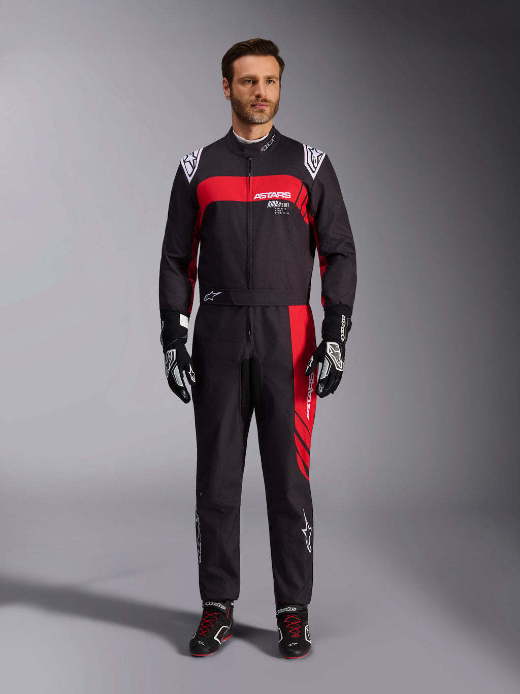 Tuta Alpinestars KMX-9 V3 Graphic 3, tuta da karting, nero e rosso, vista frontale a figura intera indossata da un modello, con pannelli grafici rossi e loghi bianchi, abbinata a guanti e scarpe da corsa neri, abbigliamento professionale per motorsport