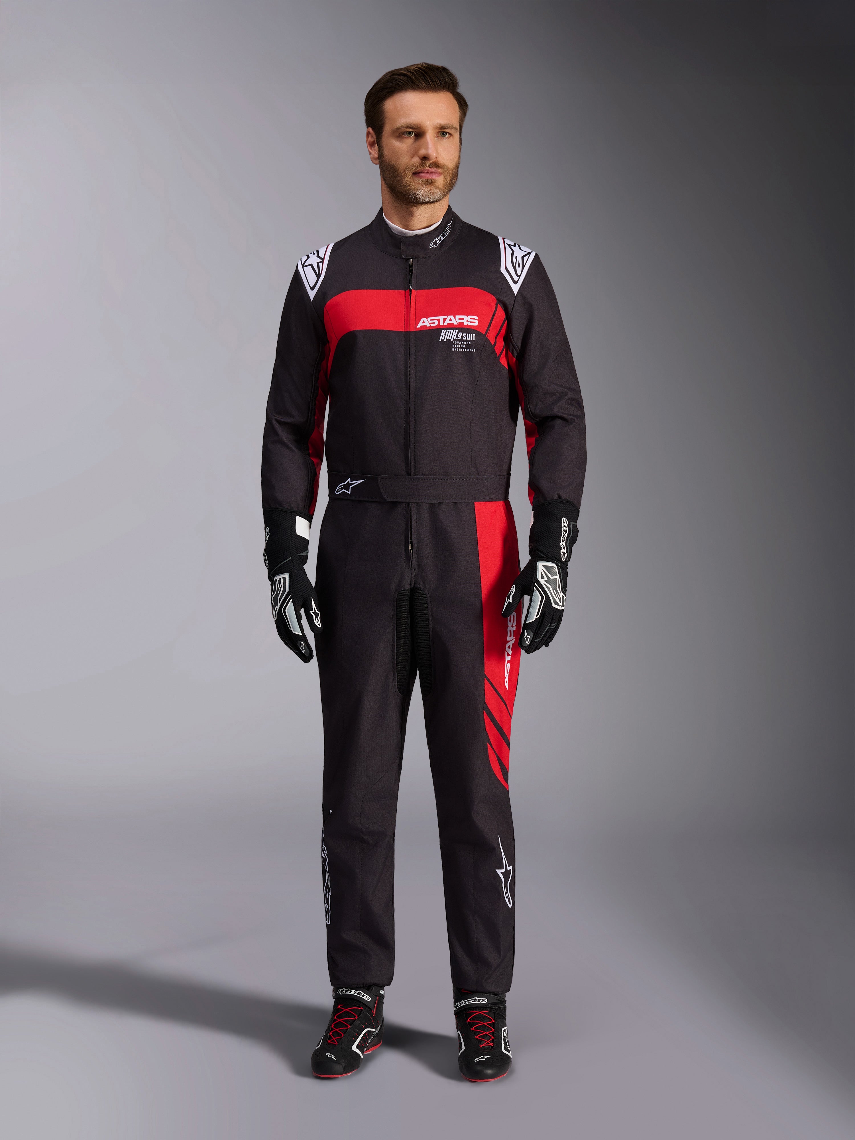 Tuta Alpinestars KMX-9 V3 Graphic 3, tuta da karting, nero e rosso, vista frontale a figura intera indossata da un modello, con pannelli grafici rossi e loghi bianchi, abbinata a guanti e scarpe da corsa neri, abbigliamento professionale per motorsport