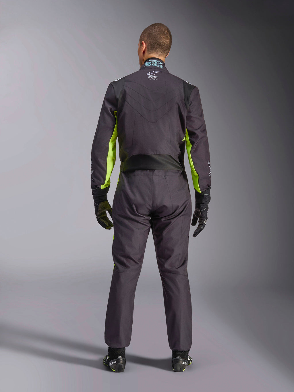 Vista posteriore a figura intera di una persona che indossa la Tuta Alpinestars KMX-9 V3 Graphic 3, tuta da karting in nero e giallo fluo, che mostra i pannelli elastici ergonomici sulla schiena e sulle spalle, abbinata a guanti da corsa e calzature neri.