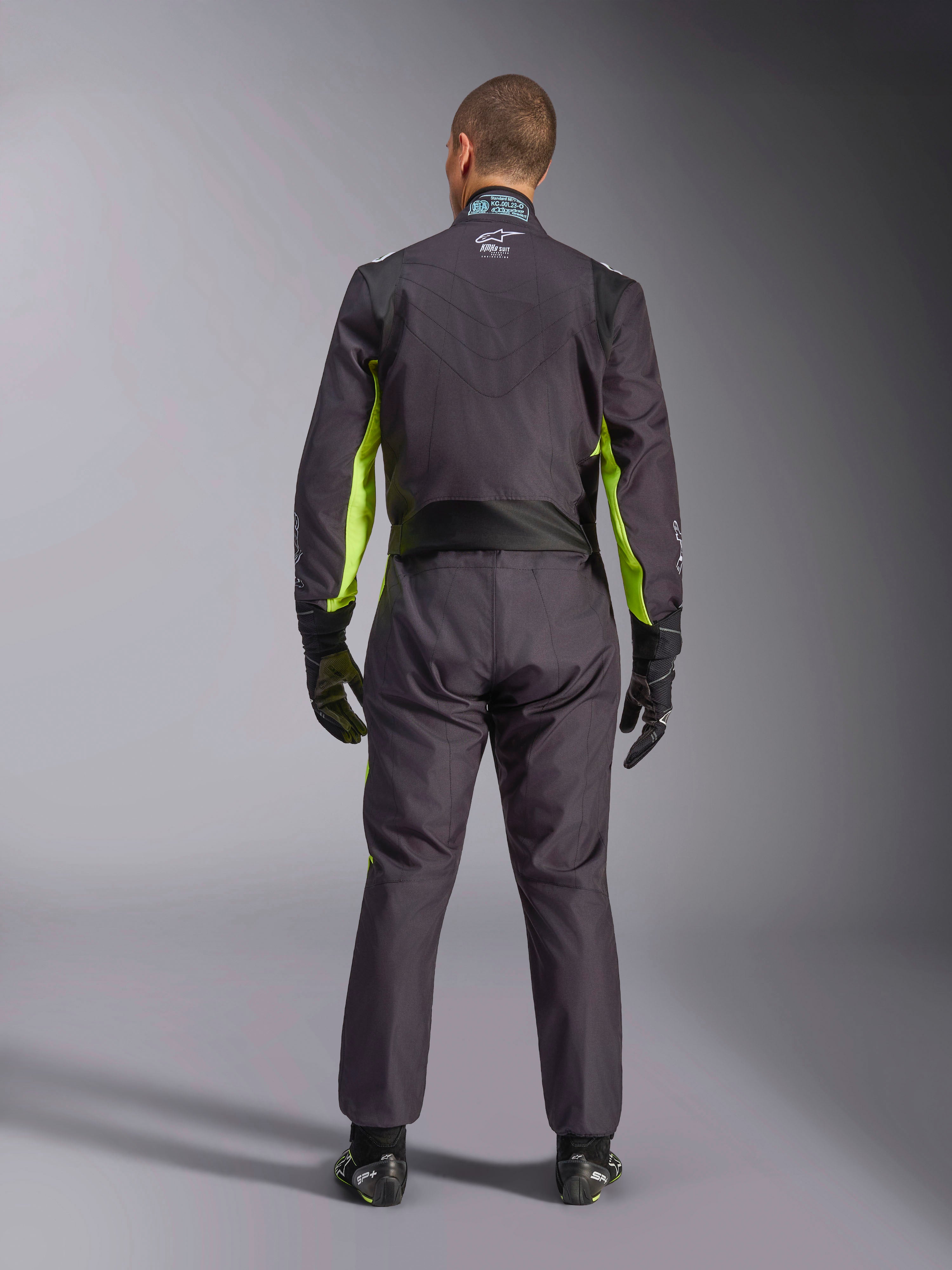 Vista posteriore a figura intera di una persona che indossa la Tuta Alpinestars KMX-9 V3 Graphic 3, tuta da karting in nero e giallo fluo, che mostra i pannelli elastici ergonomici sulla schiena e sulle spalle, abbinata a guanti da corsa e calzature neri.