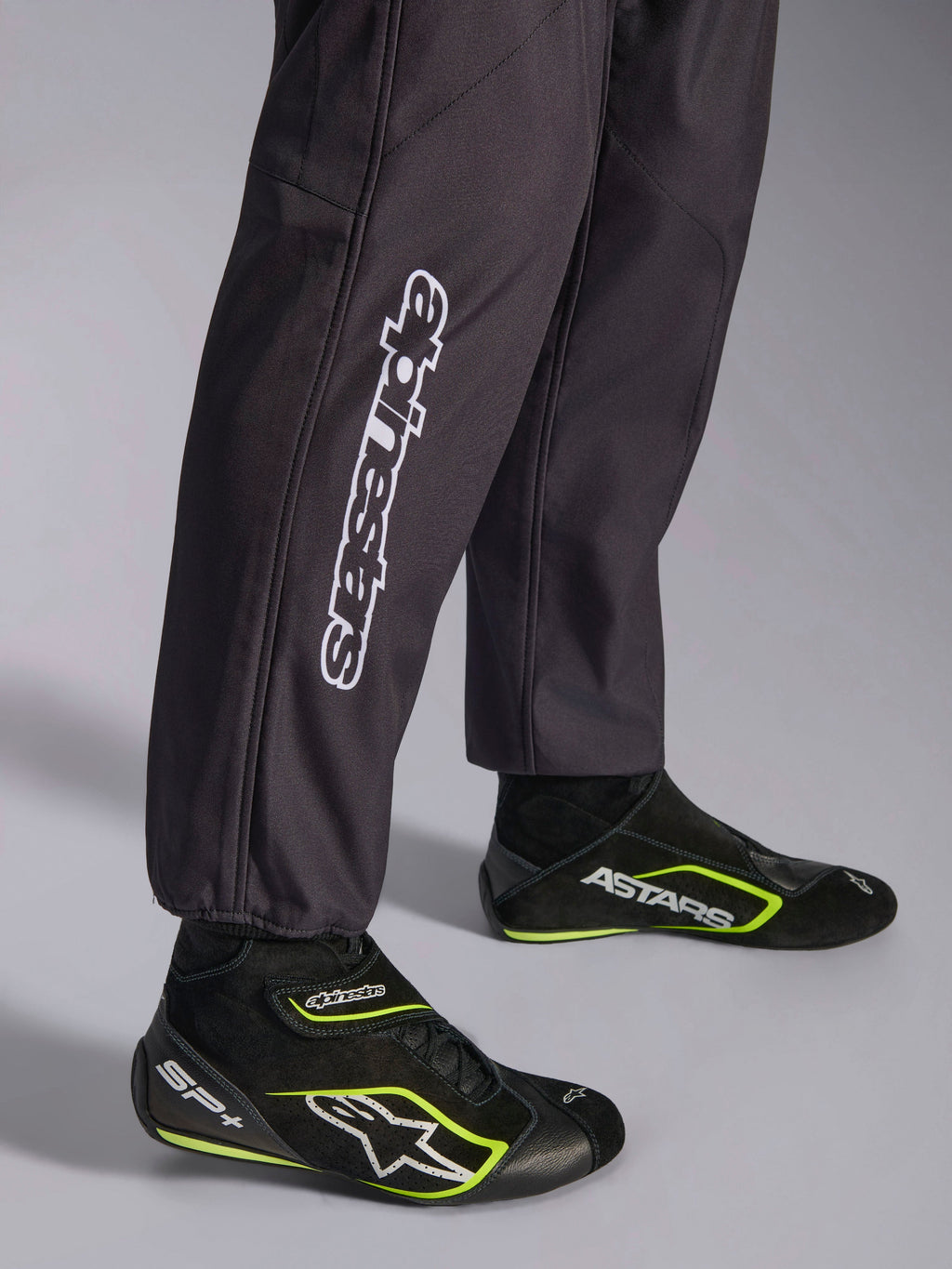 Tuta Alpinestars KMX-9 V3 Graphic 3, tuta da karting, nera, primo piano della parte inferiore delle gambe e dei polsini alle caviglie, con branding Alpinestars verticale bianco, abbinata a scarpe da karting nere e giallo fluo, dettaglio di abbigliamento professionale da karting