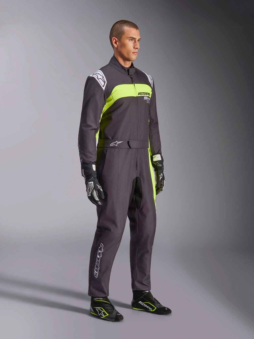 Modello con Tuta Alpinestars KMX-9 V3 Graphic 3 in nero e giallo fluo, abbigliamento professionale da karting con vivaci pannelli giallo neon e dettagli bianchi, accessoriato con guanti e scarpe da corsa coordinati, su sfondo da studio