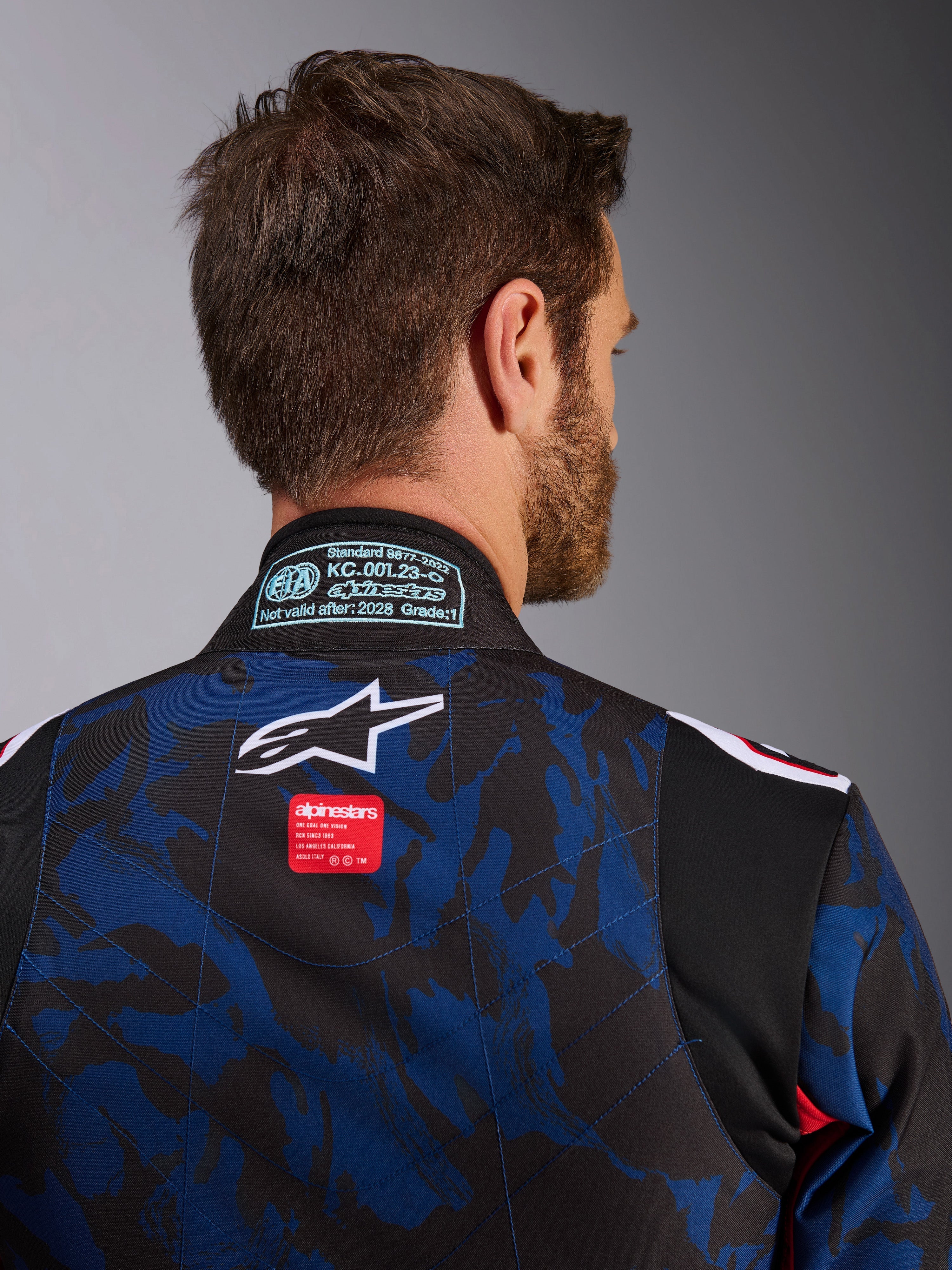 Dettaglio della tuta Alpinestars KMX-9 V3 Graphic 5, tuta da kart in Navy Red, con motivo camouflage blu e nero, logo a stella bianco ed etichetta rossa sulla parte superiore della schiena, con patch di certificazione FIA 8877-2022 sul colletto