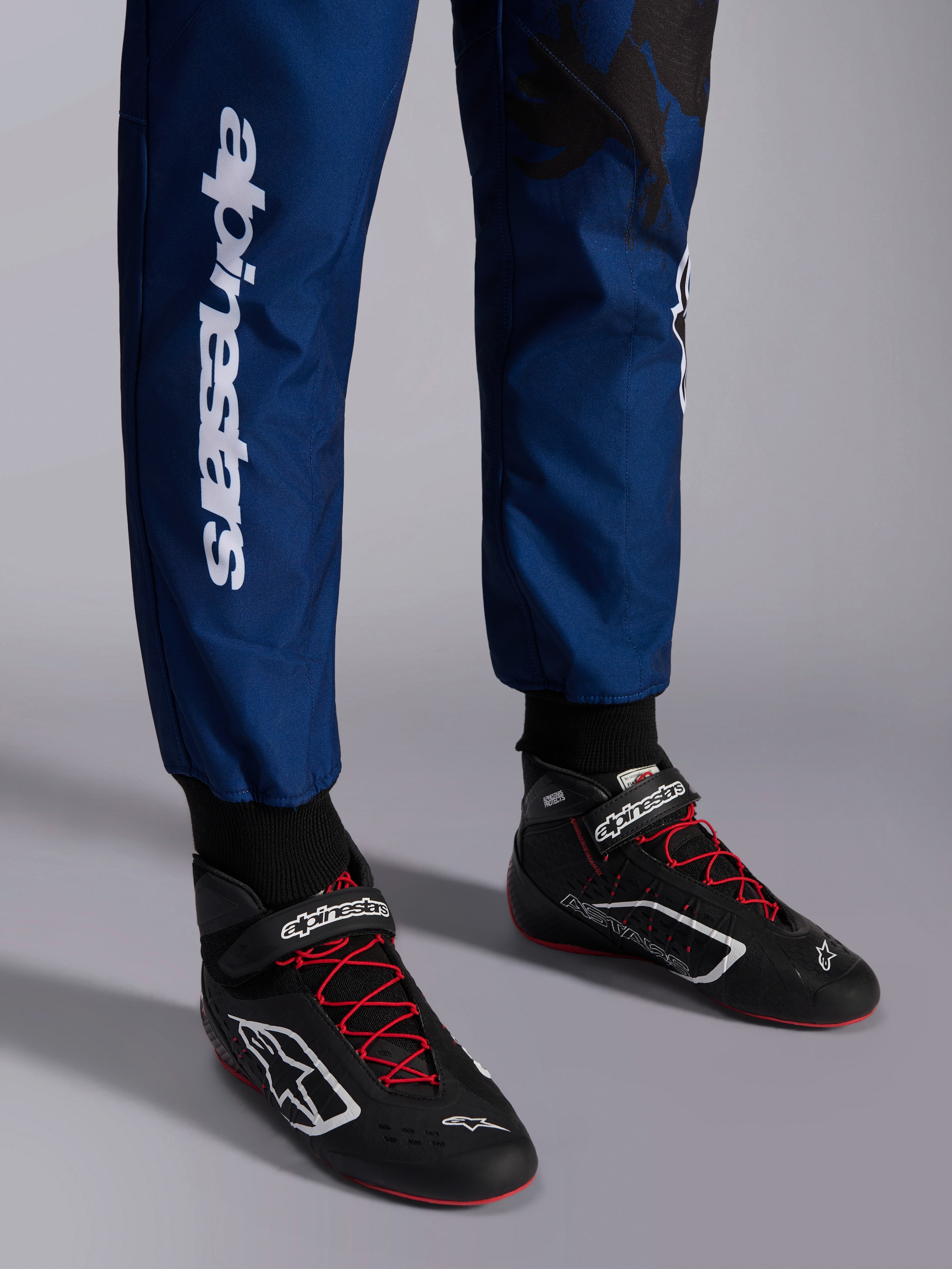 Tuta Alpinestars KMX-9 V3 Graphic 5, Navy Rosso, primo piano della parte inferiore delle gambe e scarpe da kart, tessuto blu navy con marchio Alpinestars bianco verticale, scarpe nere con lacci rossi e loghi bianchi, abbigliamento professionale da karting