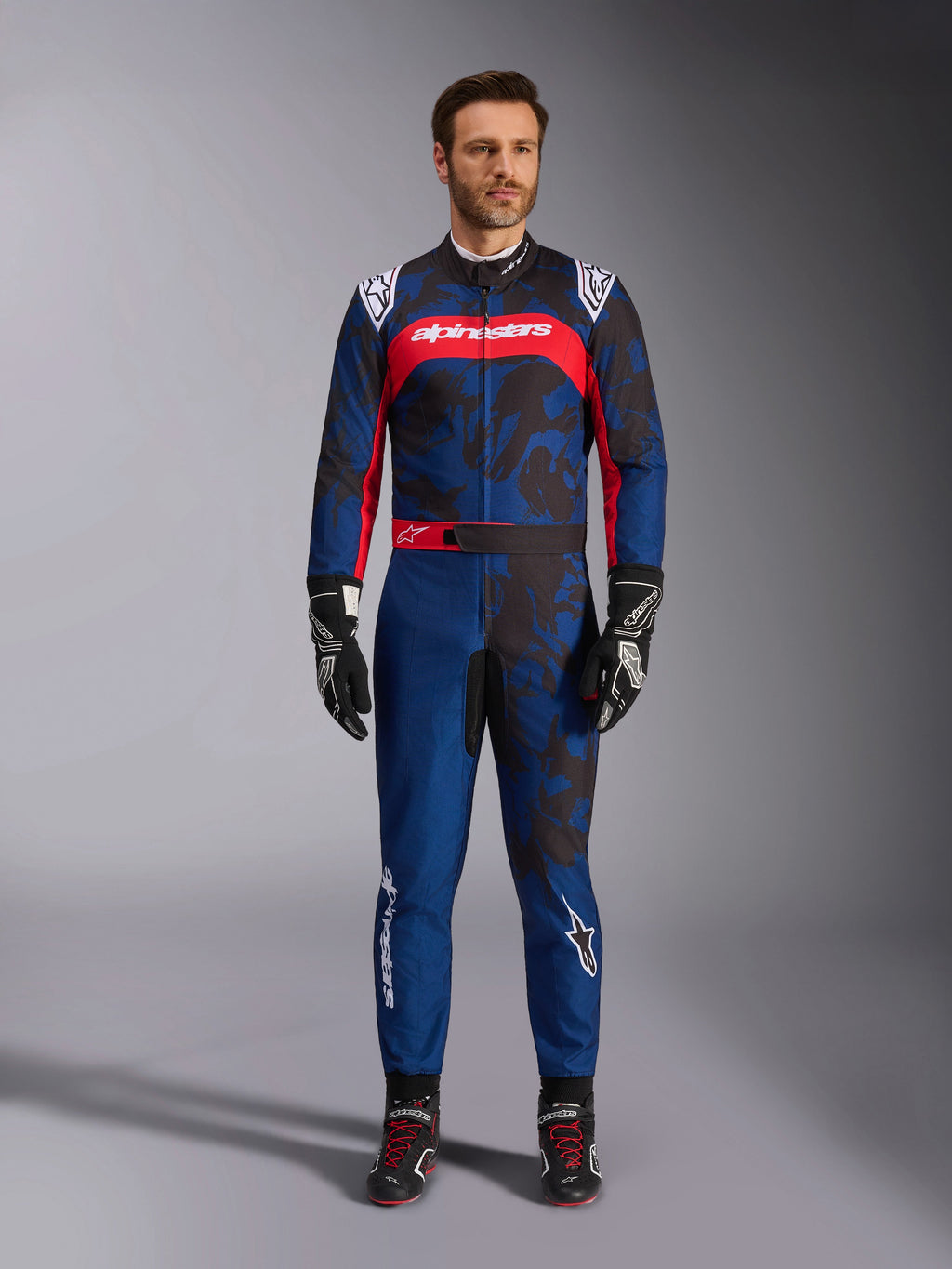 Tuta Alpinestars KMX-9 V3 Graphic 5, tuta da kart in blu navy e rosso, caratterizzata da un motivo grafico astratto in stile camo, indossata da un modello con guanti e scarpe da corsa neri, design atletico con loghi Alpinestars in evidenza su petto e gambe.