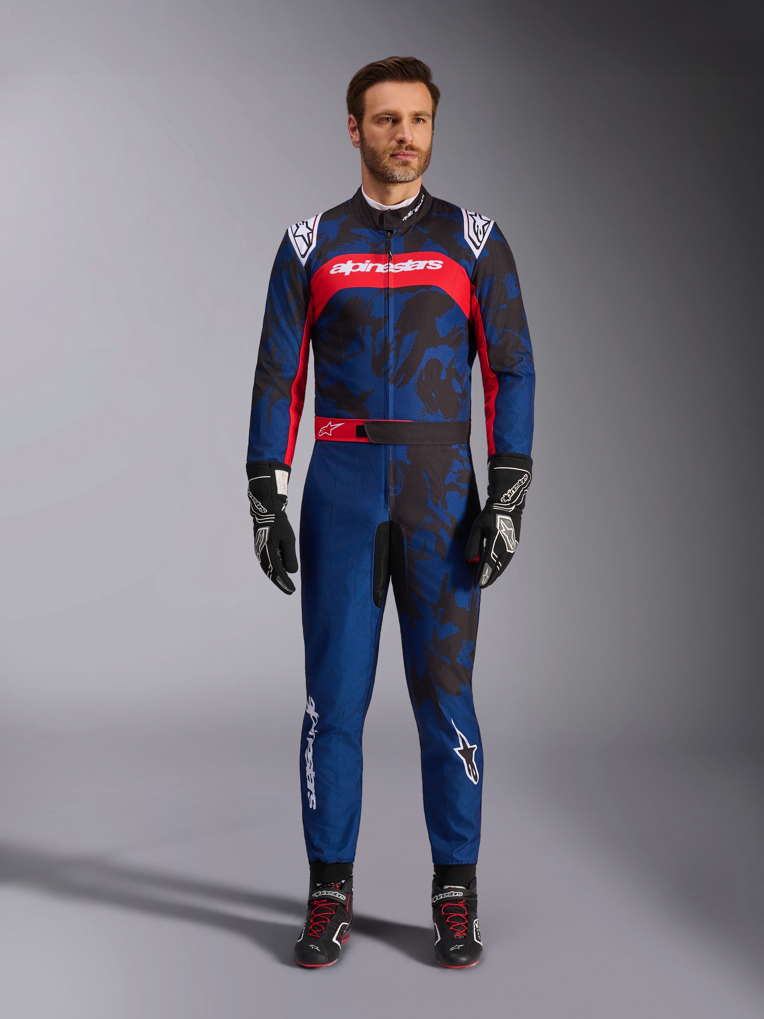 Tuta Alpinestars KMX-9 V3 Graphic 5, tuta da kart in blu navy e rosso, caratterizzata da un motivo grafico astratto in stile camo, indossata da un modello con guanti e scarpe da corsa neri, design atletico con loghi Alpinestars in evidenza su petto e gambe.