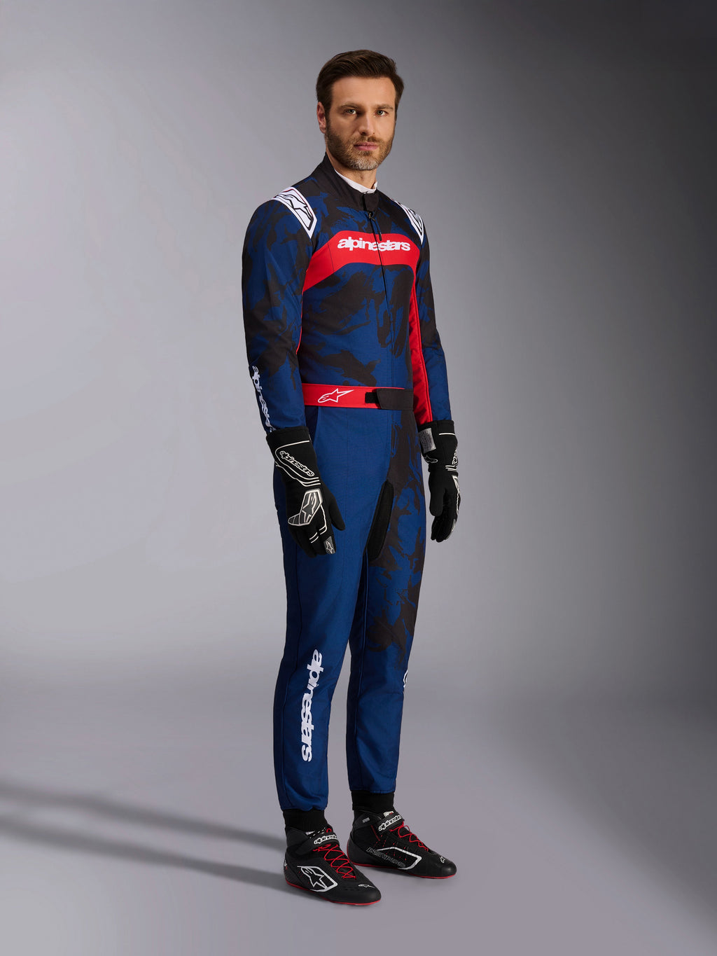 Modello che indossa la tuta Alpinestars KMX-9 V3 Graphic 5 in Navy Red, inquadratura a figura intera completa di guanti da kart neri e scarpe da corsa nere, tuta blu navy con grafiche scure stile camouflage e un pannello rosso sul petto con logo bianco, abbigliamento protettivo professionale per il karting
