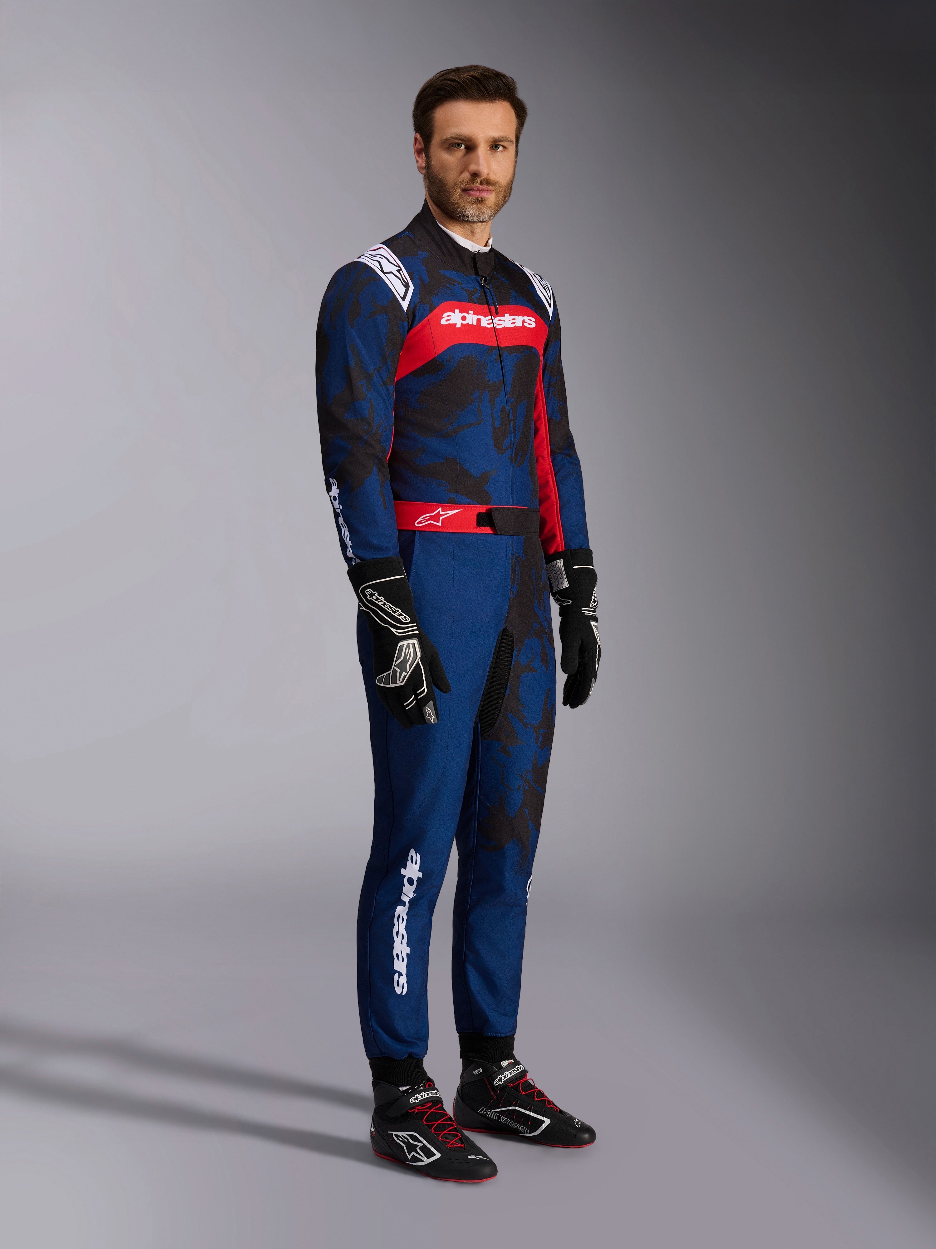 Modello che indossa la tuta Alpinestars KMX-9 V3 Graphic 5 in Navy Red, inquadratura a figura intera completa di guanti da kart neri e scarpe da corsa nere, tuta blu navy con grafiche scure stile camouflage e un pannello rosso sul petto con logo bianco, abbigliamento protettivo professionale per il karting