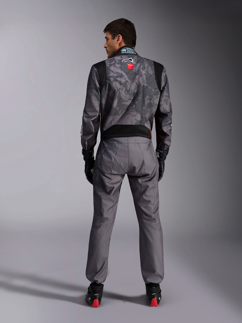 Alpinestars KMX-9 V3 Graphic 5 Suit, tuta da karting, motivo camouflage grigio scuro e nero sulla parte superiore del corpo, vista posteriore che mostra i pannelli elasticizzati e il marchio Alpinestars, indossata con guanti neri e scarpe da corsa nere con dettagli rossi, abbigliamento professionale per competizioni motoristiche