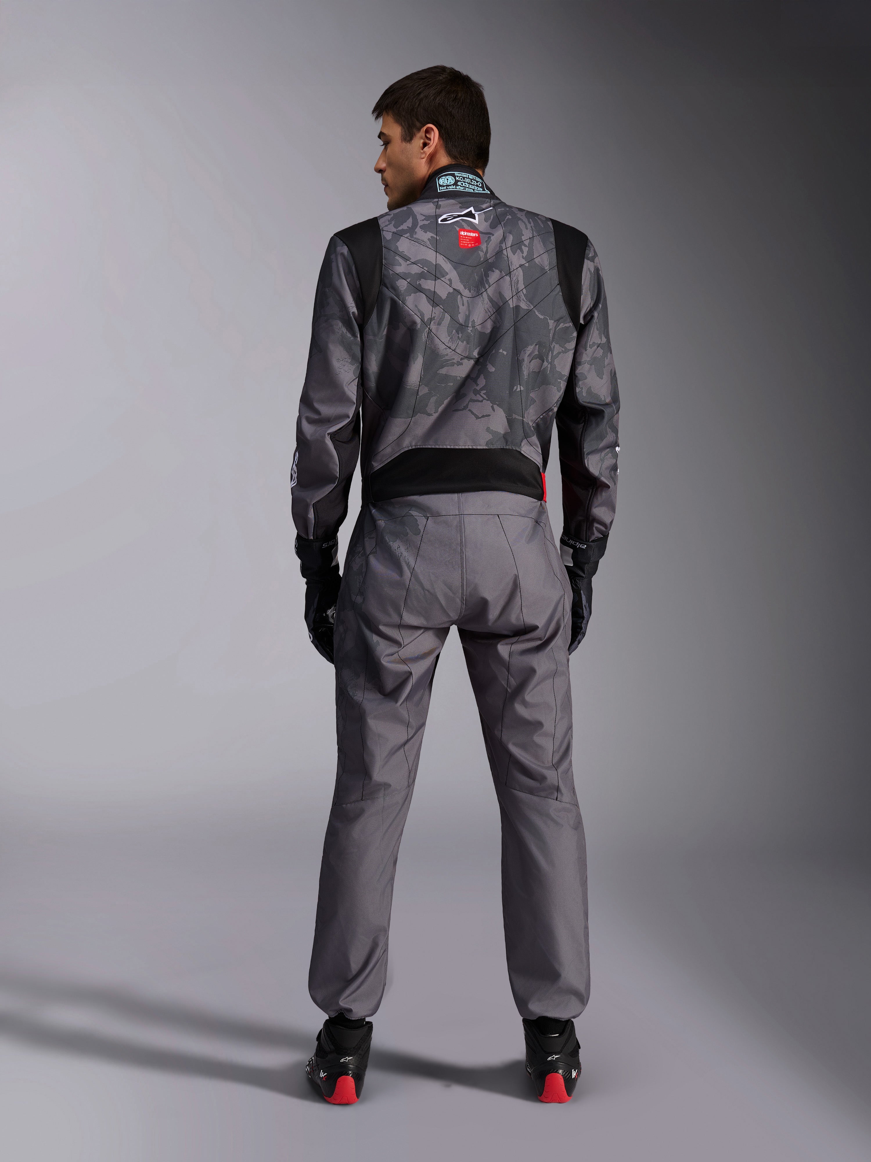 Alpinestars KMX-9 V3 Graphic 5 Suit, tuta da karting, motivo camouflage grigio scuro e nero sulla parte superiore del corpo, vista posteriore che mostra i pannelli elasticizzati e il marchio Alpinestars, indossata con guanti neri e scarpe da corsa nere con dettagli rossi, abbigliamento professionale per competizioni motoristiche