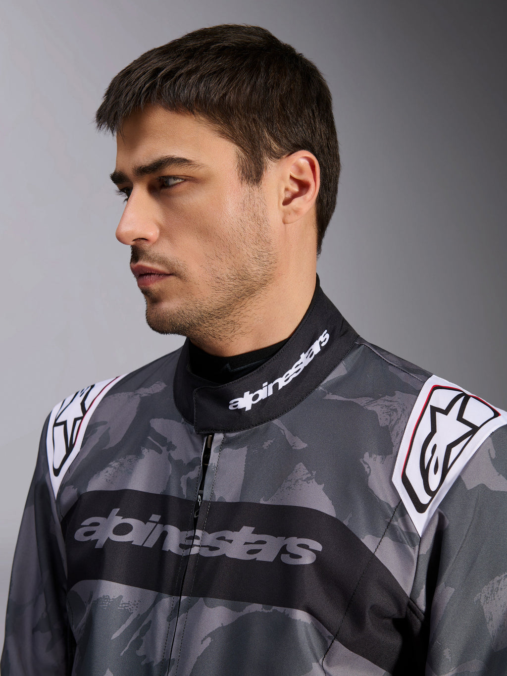 Tuta Alpinestars KMX-9 V3 Graphic 5, tuta da kart, motivo mimetico grigio scuro e nero, primo piano del colletto e delle spalle con loghi bianchi, abbigliamento da corsa professionale