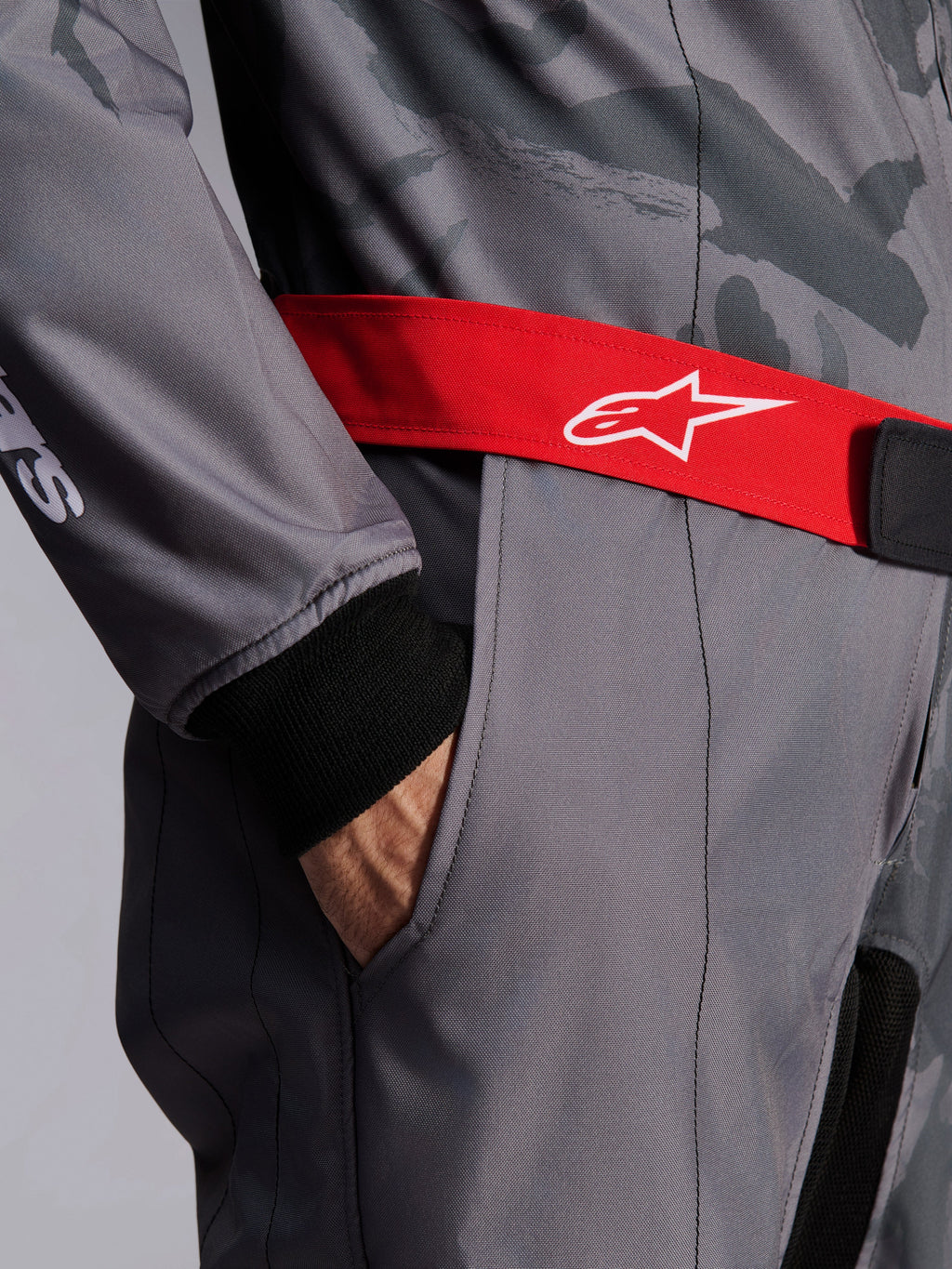 Alpinestars KMX-9 V3 Graphic 5 Suit, tuta da karting, grigio scuro e nero con cintura rossa, primo piano con cintura regolabile rossa con logo bianco, mano nella tasca sulla coscia e polsino in maglia nera su tessuto grigio con motivo grafico