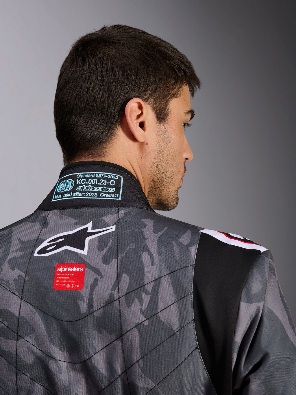 Tuta Alpinestars KMX-9 V3 Graphic 5, tuta da kart, motivo mimetico grigio scuro e nero, primo piano della parte superiore della schiena che mostra il logo Alpinestars, etichetta rossa del brand ed etichetta di omologazione FIA sul colletto.