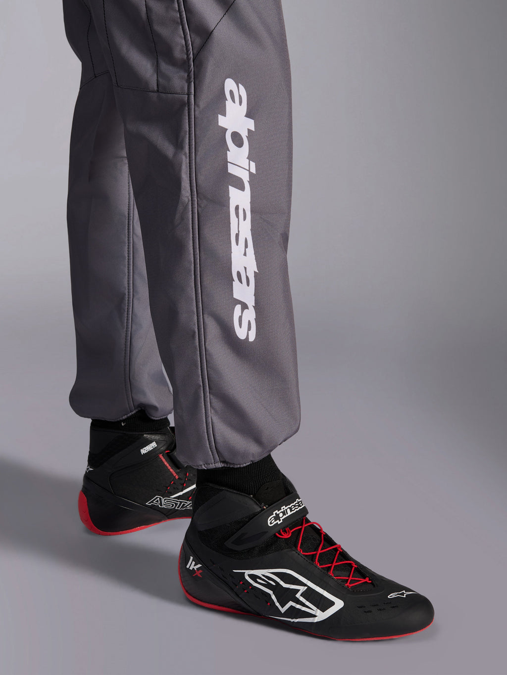 Tuta Alpinestars KMX-9 V3 Graphic 5, tuta da kart, grigio scuro, primo piano della parte inferiore della gamba con logo Alpinestars verticale bianco, indossata con scarpe da corsa nere e rosse, materiale resistente per competizioni di kart