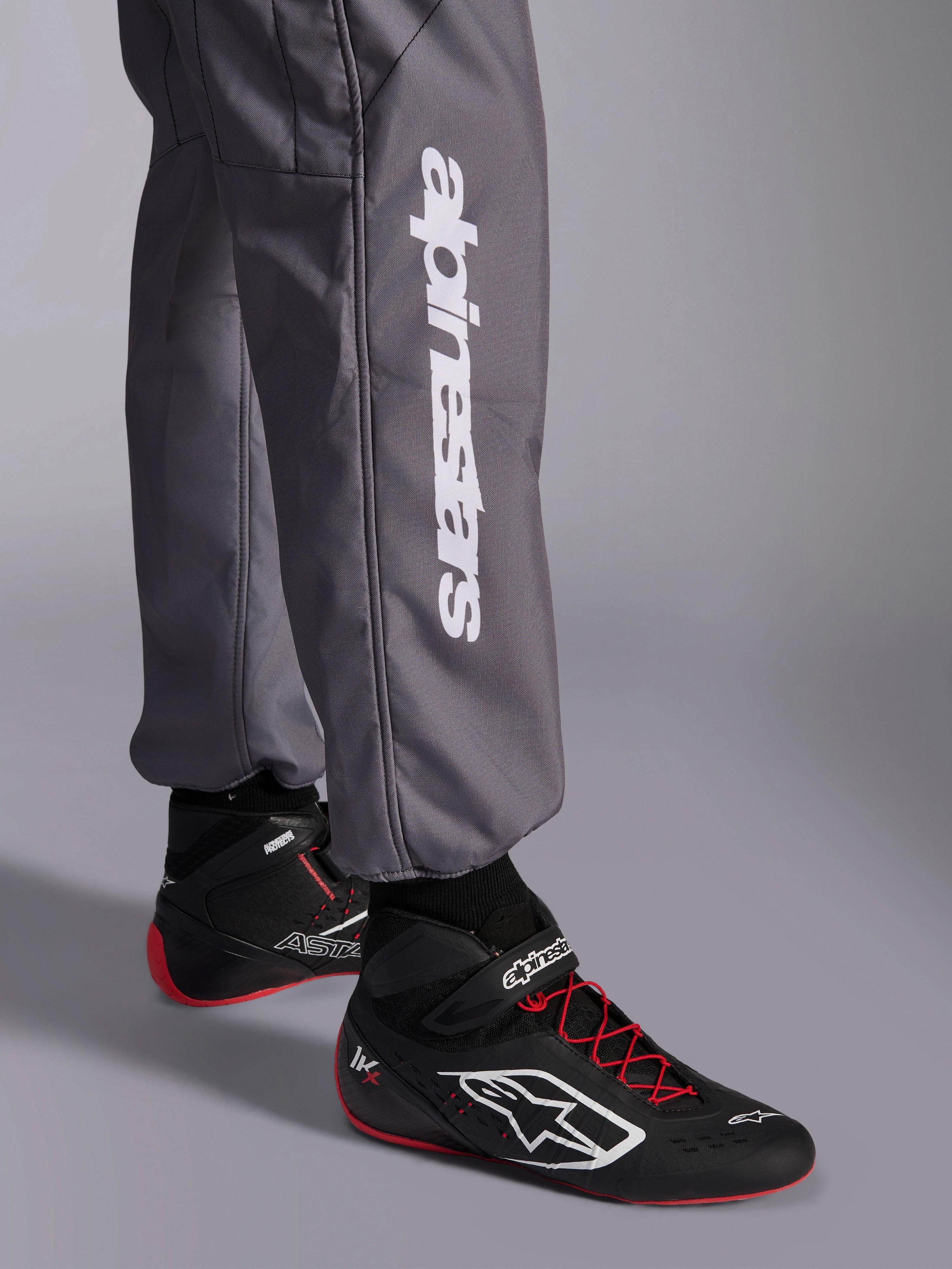 Tuta Alpinestars KMX-9 V3 Graphic 5, tuta da kart, grigio scuro, primo piano della parte inferiore della gamba con logo Alpinestars verticale bianco, indossata con scarpe da corsa nere e rosse, materiale resistente per competizioni di kart