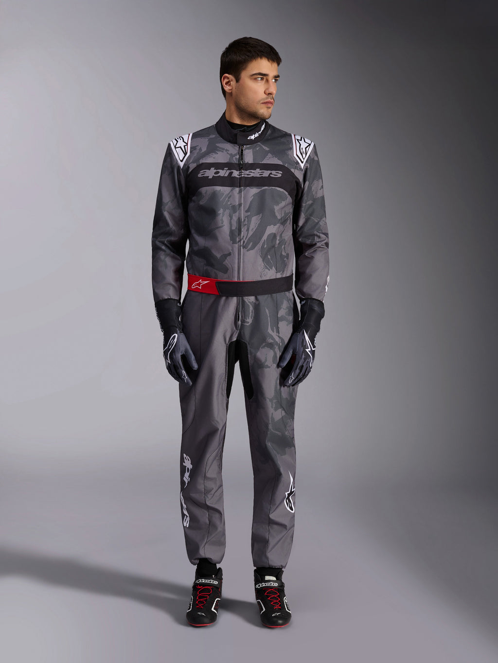 Tuta Alpinestars KMX-9 V3 Graphic 5, tuta da karting, grigio scuro e nero con grafiche in stile camouflage, con fascia nera sul petto con logo bianco e dettaglio rosso sulla cintura, indossata con guanti e scarpe da corsa coordinati, abbigliamento professionale per motorsport