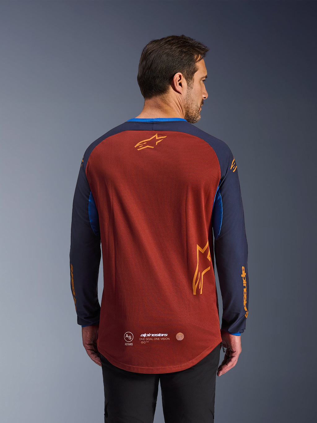 Alpinestars Maxdura Dual Jersey in Rust Brown e Mood Indigo, vista dal retro, caratterizzata da busto in mesh traspirante, maniche blu scuro con logo Alpinestars arancione e orlo posteriore allungato per una maggiore copertura.