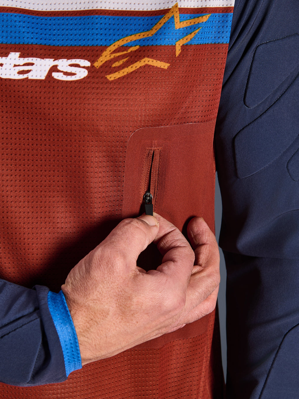 Primo piano della maglia Alpinestars Maxdura Dual, busto in mesh Rust Brown con tasca sul petto senza cuciture con cerniera, maniche Mood Indigo con imbottitura integrata, dettaglio tecnico di maglia da moto adventure