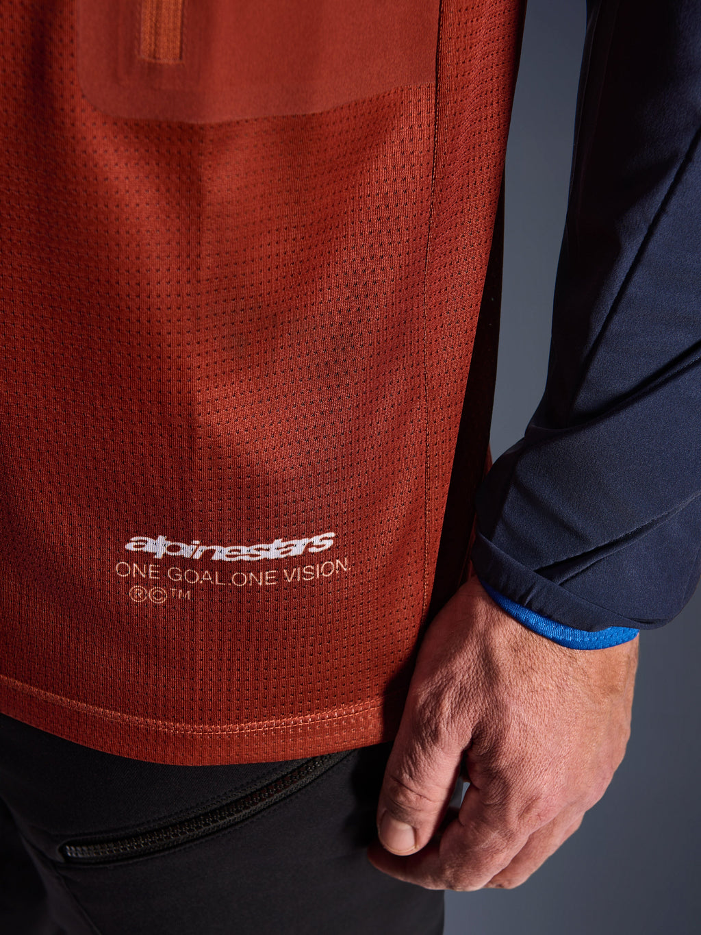 Alpinestars Maxdura Dual Jersey, maglia da moto adventure, marrone ruggine e blu Mood Indigo, vista ravvicinata che mostra il tessuto a rete del busto e la manica in tessuto blu scuro, logo Alpinestars 'One Goal One Vision' bianco sull'orlo inferiore