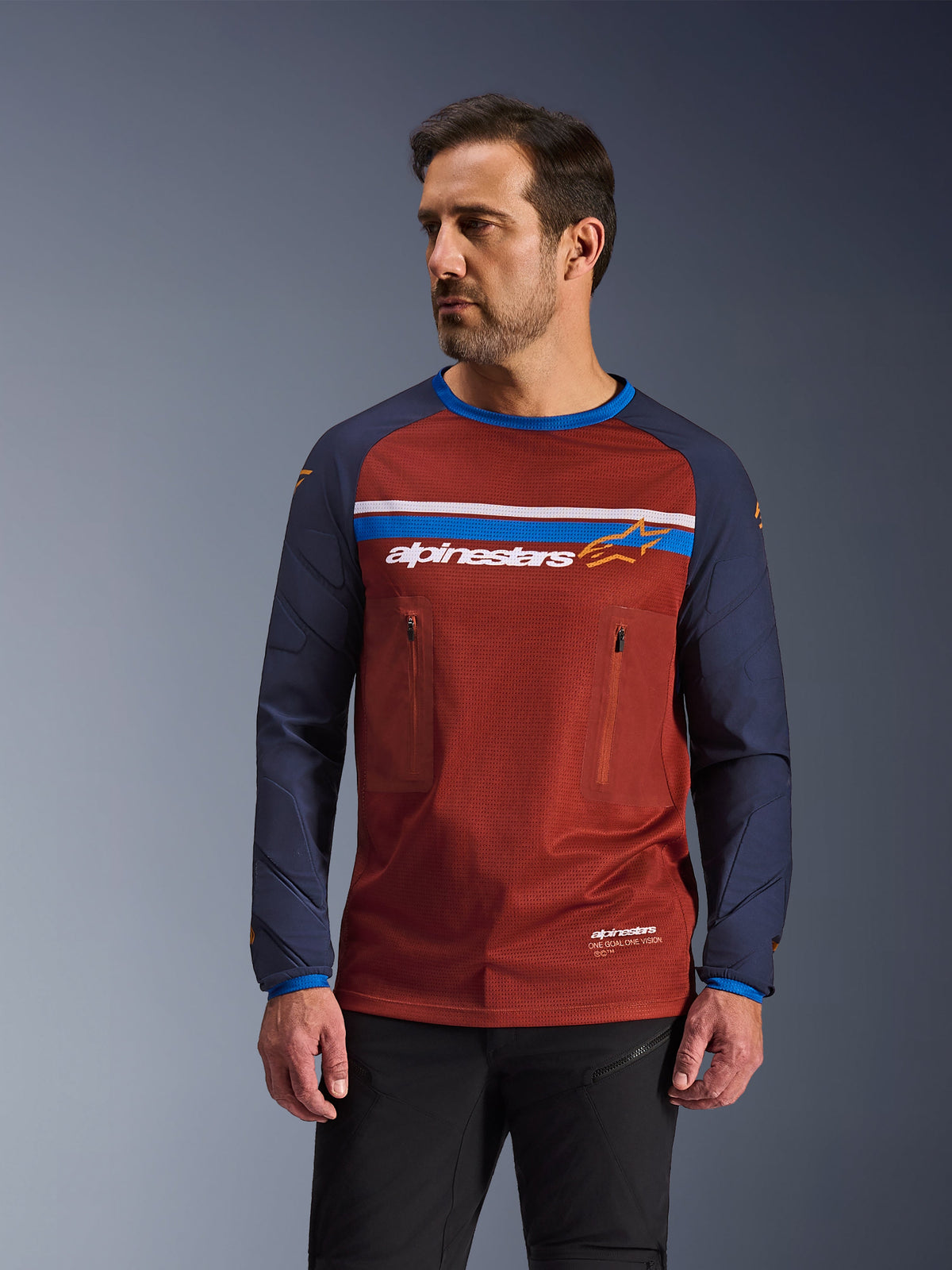 Modello che indossa Alpinestars Maxdura Dual Jersey, maglia da moto adventure marrone ruggine e blu navy, con doppie tasche sul petto con cerniera, striscia orizzontale sul petto con marchio, busto in rete tecnica e maniche in tessuto resistente.