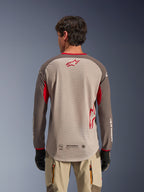 Vista posteriore di un uomo che indossa la maglia Alpinestars Maxdura Dual in Aluminum Walnut, maglia da moto adventure con busto in rete sabbia e logo a stella rosso, maniche marrone scuro con accenti rossi, abbinata a pantaloni da guida beige e guanti neri, progettata per ventilazione off-road ad alte prestazioni.