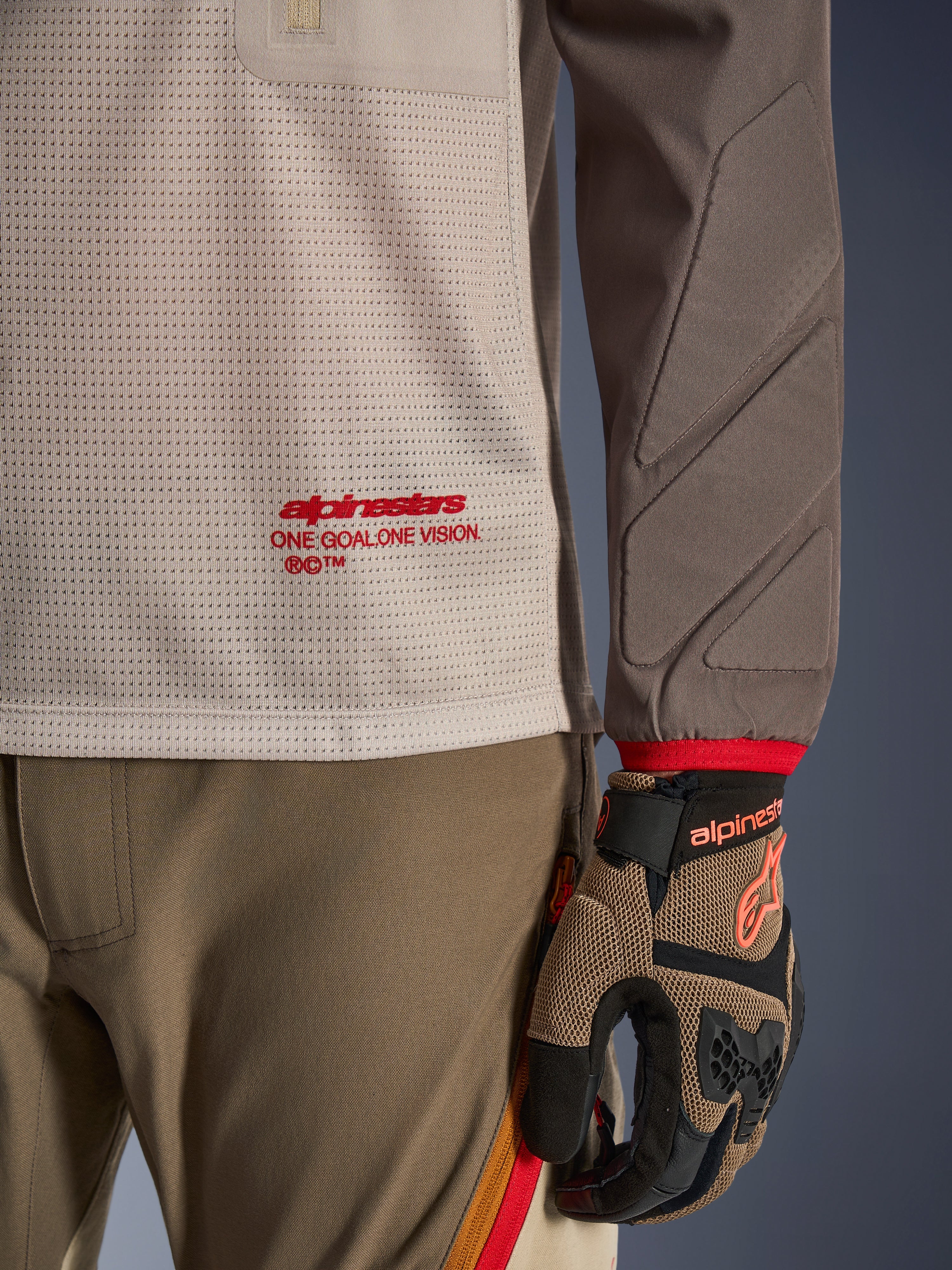 Alpinestars Maxdura Dual Jersey in Aluminum Walnut, vista dettagliata che mostra il busto in rete ventilata con logo rosso, manica resistente all'abrasione con imbottitura integrata, indossata con pantaloni e guanti da moto adventure coordinati