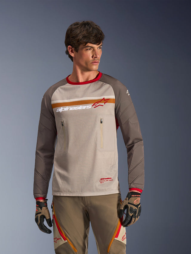 Alpinestars Maxdura Dual Jersey in Aluminum Walnut, maglia moto adventure con busto in rete traspirante, maniche marrone noce resistenti all'abrasione e tasche sul petto con cerniera, indossata da un modello con equipaggiamento off-road coordinato.