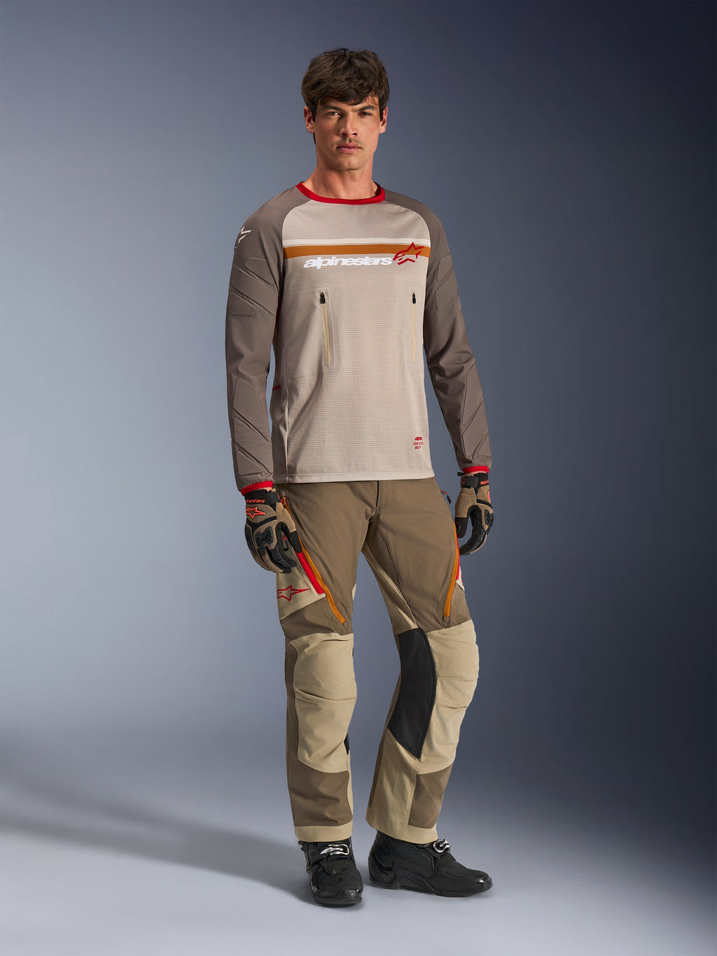 Alpinestars Maxdura Dual Jersey in Aluminum Walnut, maglia con busto in rete grigia, maniche marroni e polsini rossi, tasche sul petto con zip, indossata con abbigliamento tecnico coordinato per guida adventure.