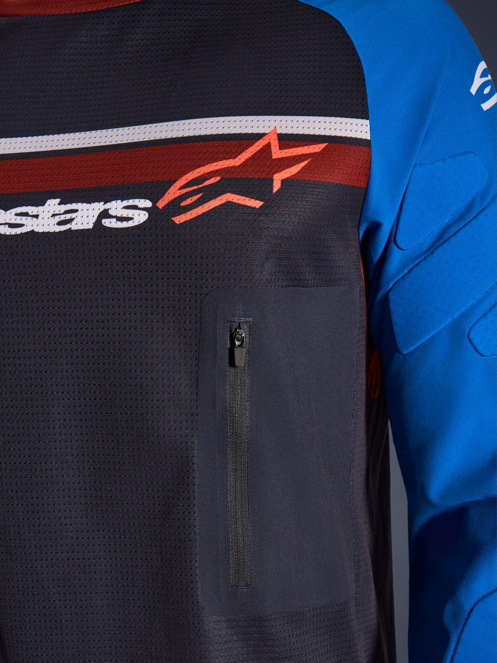 Alpinestars Maxdura Dual Jersey, primo piano di petto e manica, Deep Gray e Royal Blue, con tasca sul petto con cerniera, busto in mesh traspirante e imbottitura integrata sulle maniche, logo Alpinestars su strisce rosse e bianche.
