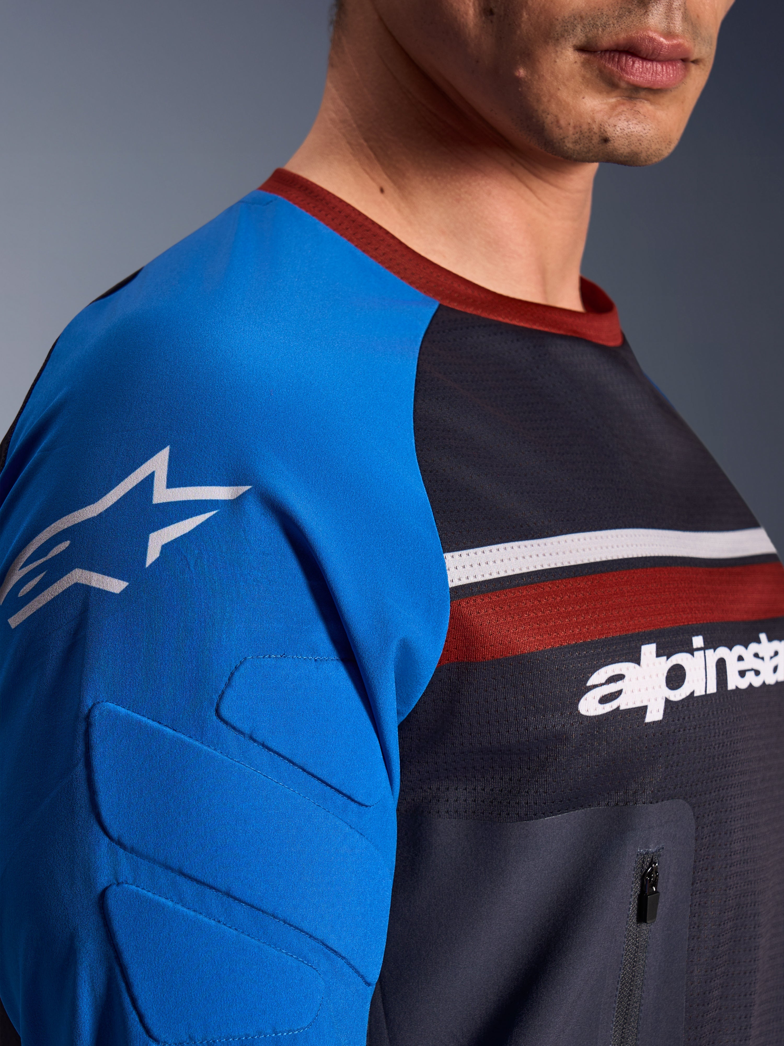 Alpinestars Maxdura Dual Jersey, maglia da moto, grigio scuro e blu reale, vista ravvicinata che mostra la tasca sul petto con cerniera, il tessuto in rete sul busto e l'imbottitura integrata sulla manica con logo a stella
