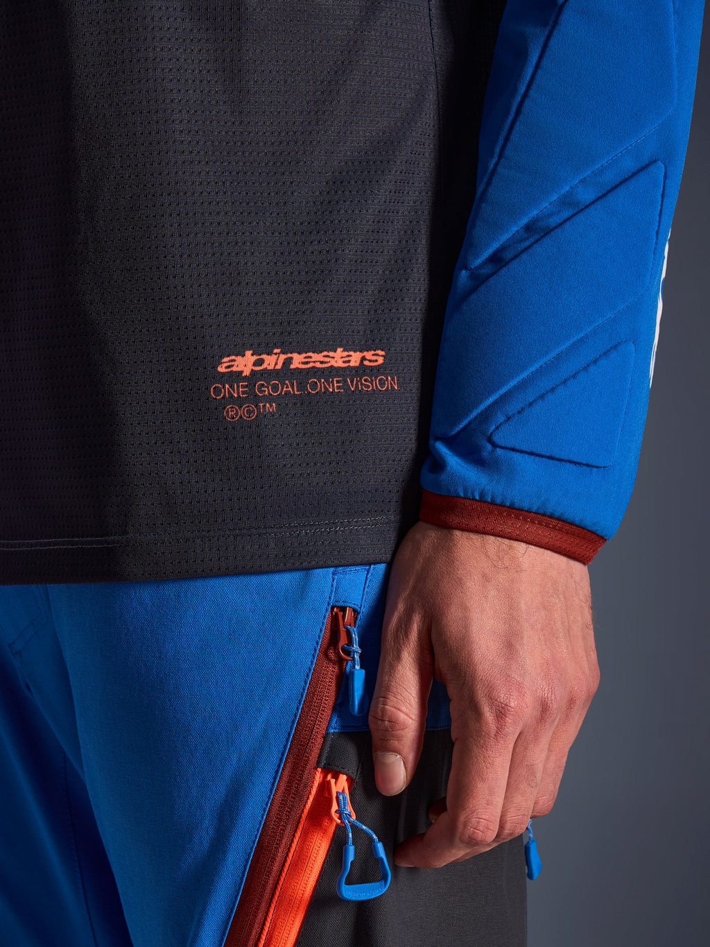 Primo piano della maglia Alpinestars Maxdura Dual in grigio scuro e blu reale, con logo arancione e scritta 'One Goal One Vision' sul busto in rete, manica imbottita con pannelli geometrici rinforzati, dettaglio tecnico per guida adventure